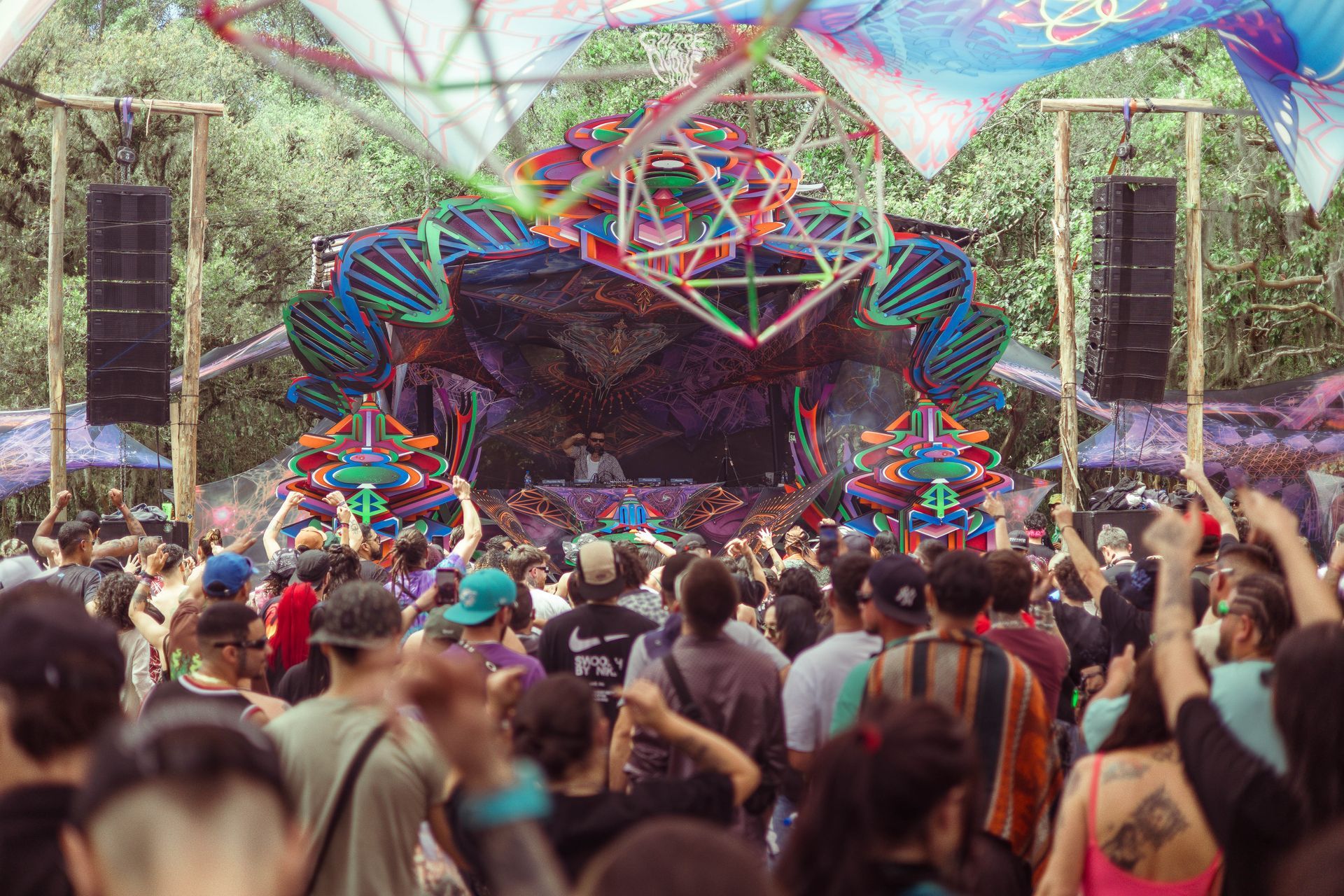 Festival de música ao ar livre lotado, com palco geométrico colorido e pessoas de frente para a cabine do DJ.
