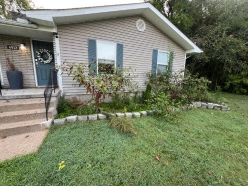 Front yard of a house with beige siding, blue shutters, a blue front door, and a small flower bed.