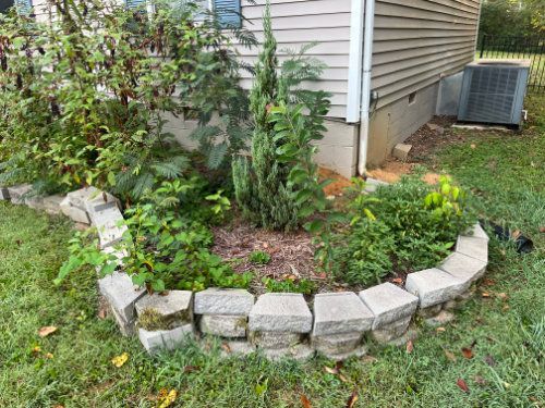 Curved stone flower bed with green plants beside a house and air conditioner unit
