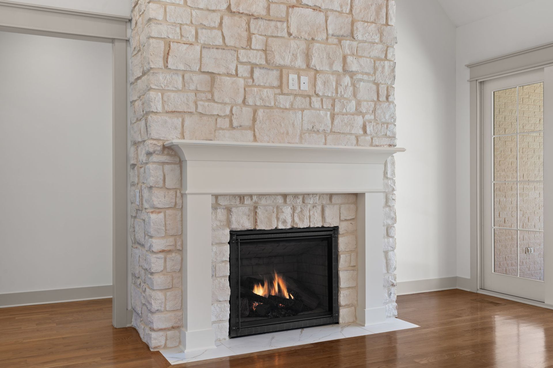 Stone fireplace with white mantel in a bright empty room with wood floors.
