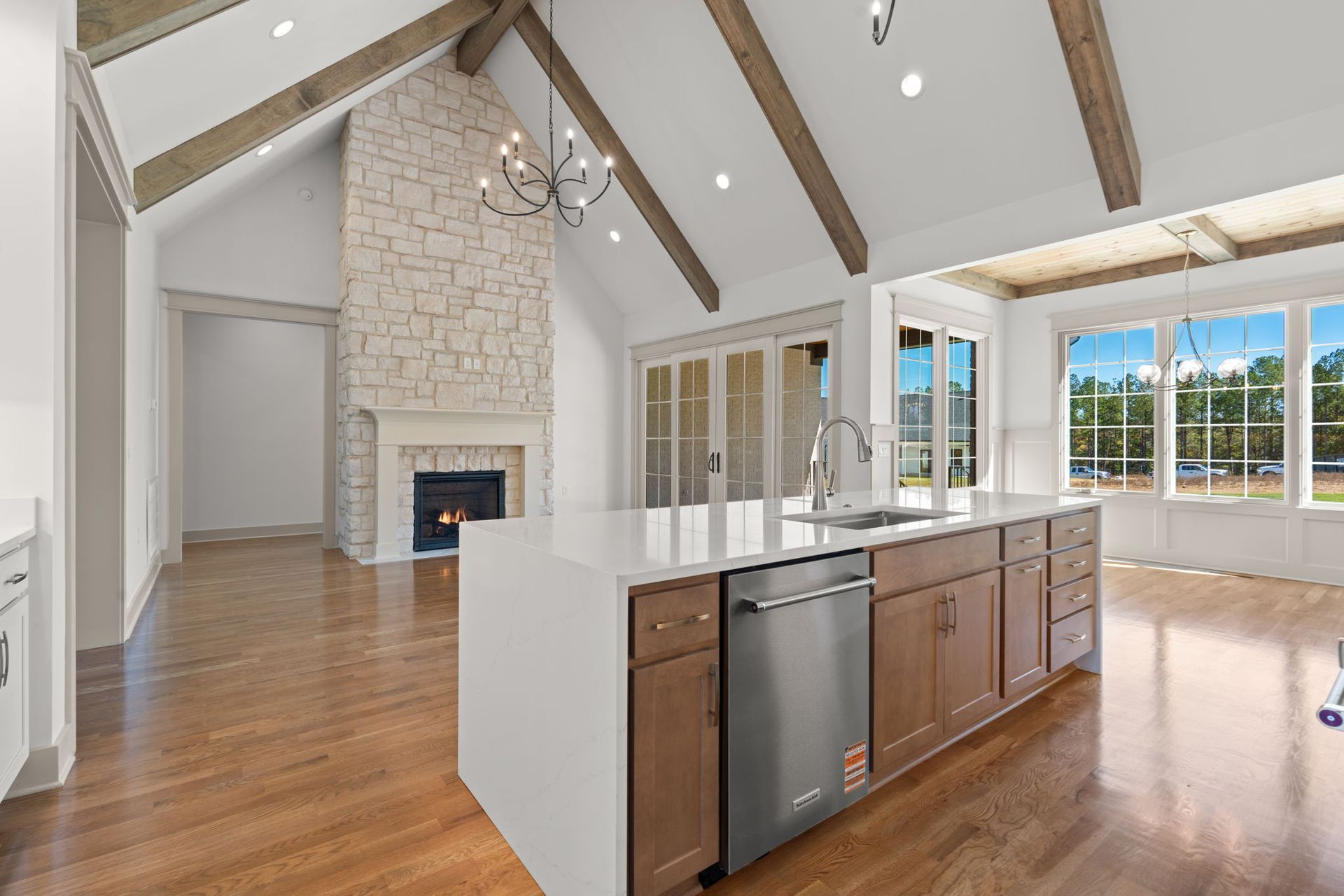 Bright open-concept kitchen and living room with wood floors, stone fireplace, island, and large windows