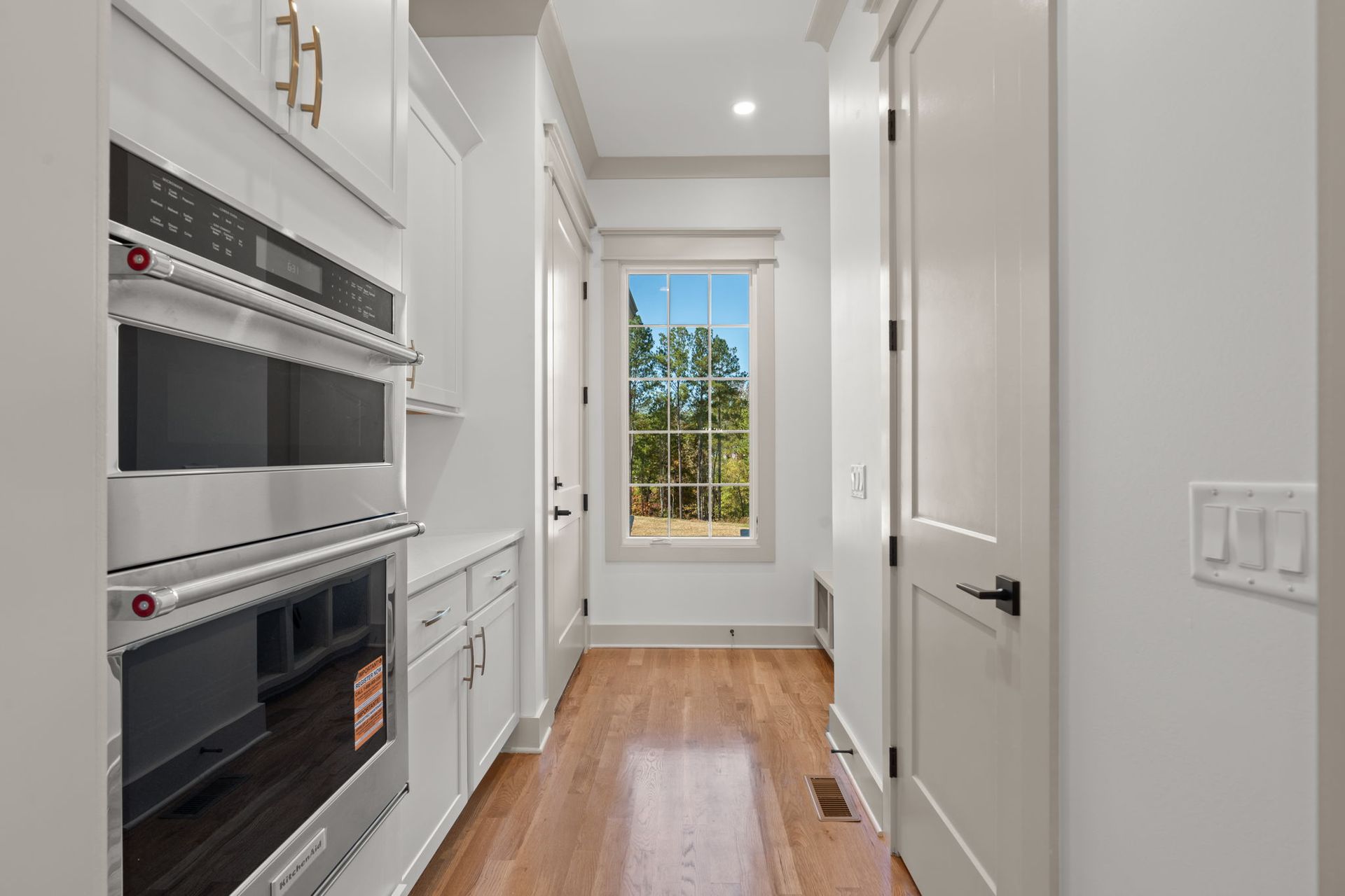 Narrow white galley kitchen with built-in ovens, wood floor, and a window at the end