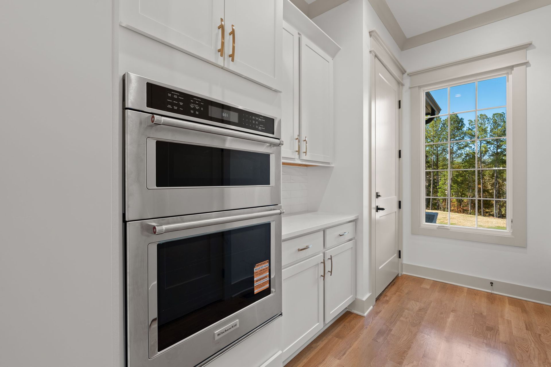 Bright white kitchen with built-in stainless double ovens, cabinets, hardwood floor, and a window.