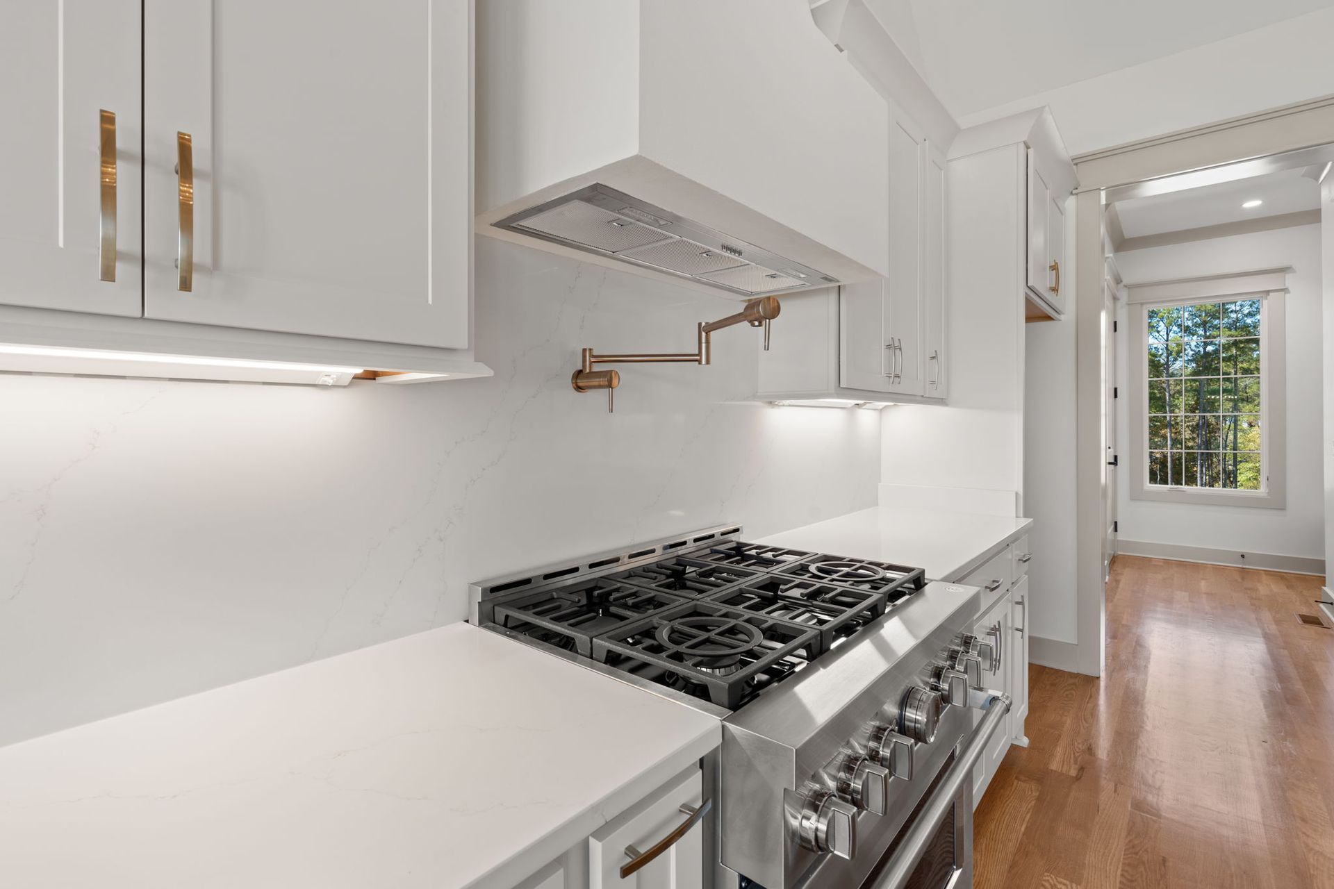 Bright white kitchen with stainless steel stove, cabinets, and hardwood floor hallway leading to a window.