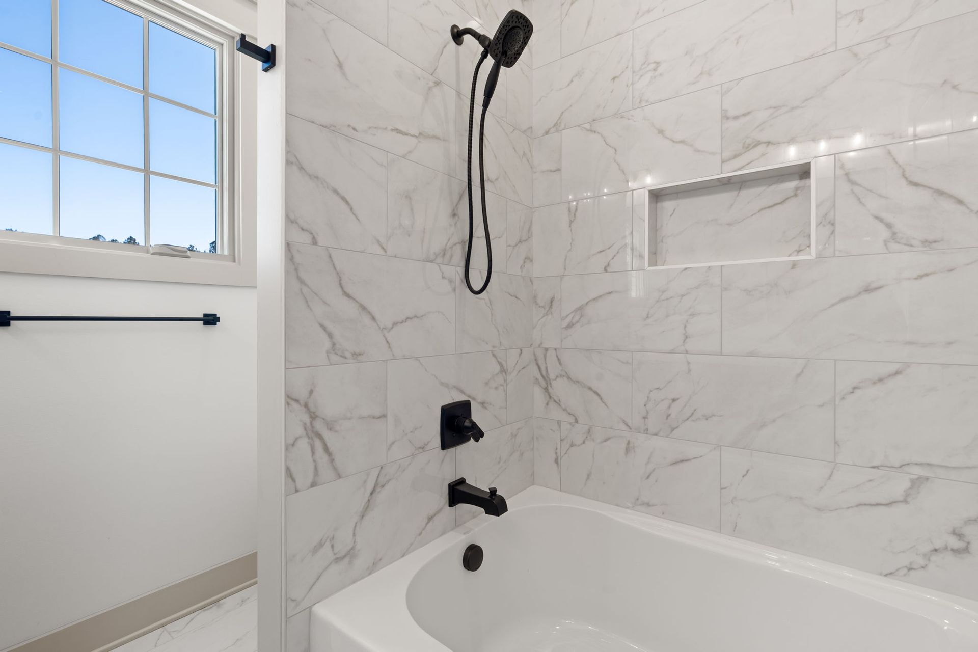 Modern white marble bathroom with bathtub, handheld shower, and frosted window.