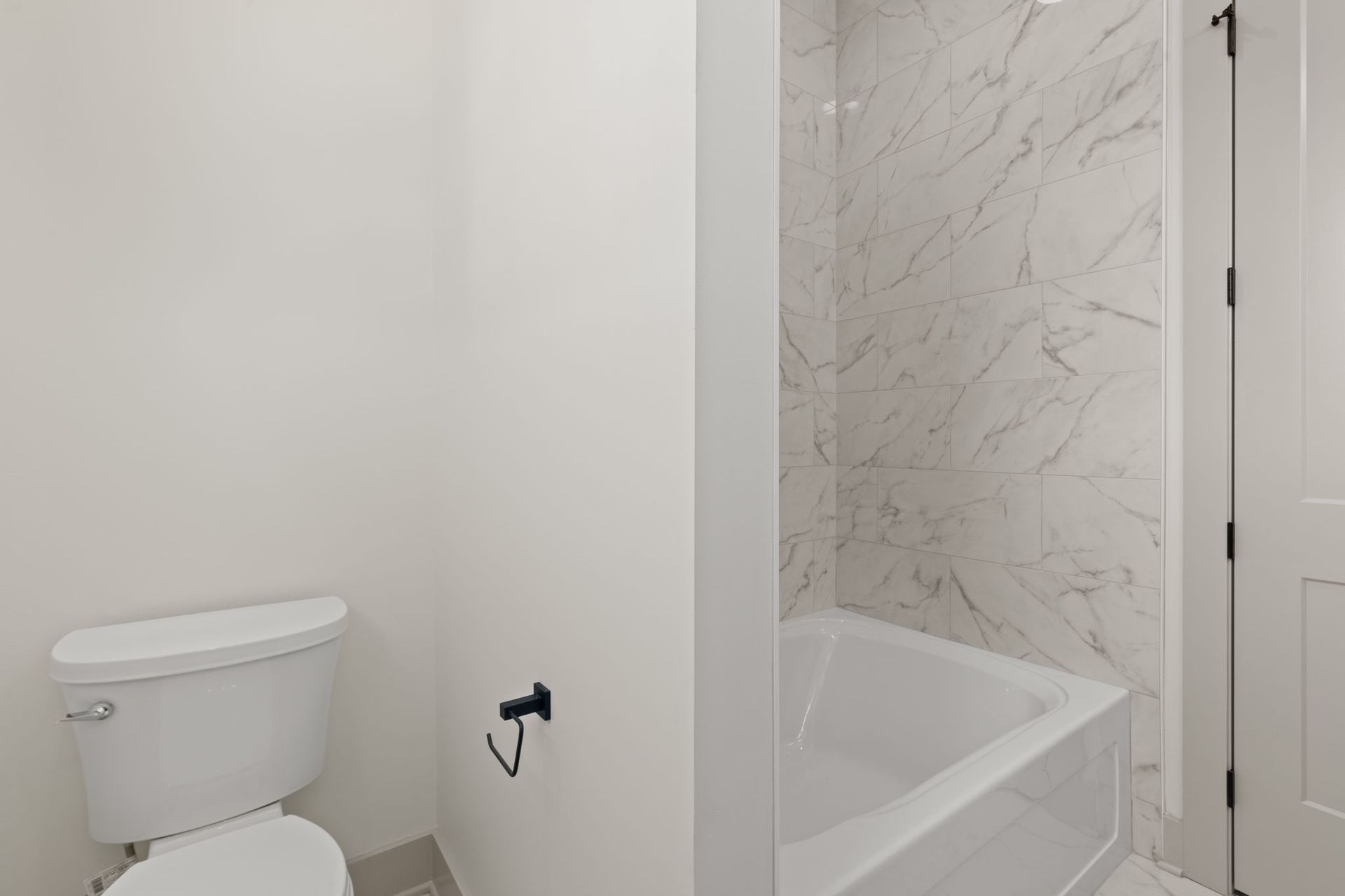 Small bathroom with toilet, bathtub, white walls, and marble-pattern shower surround.