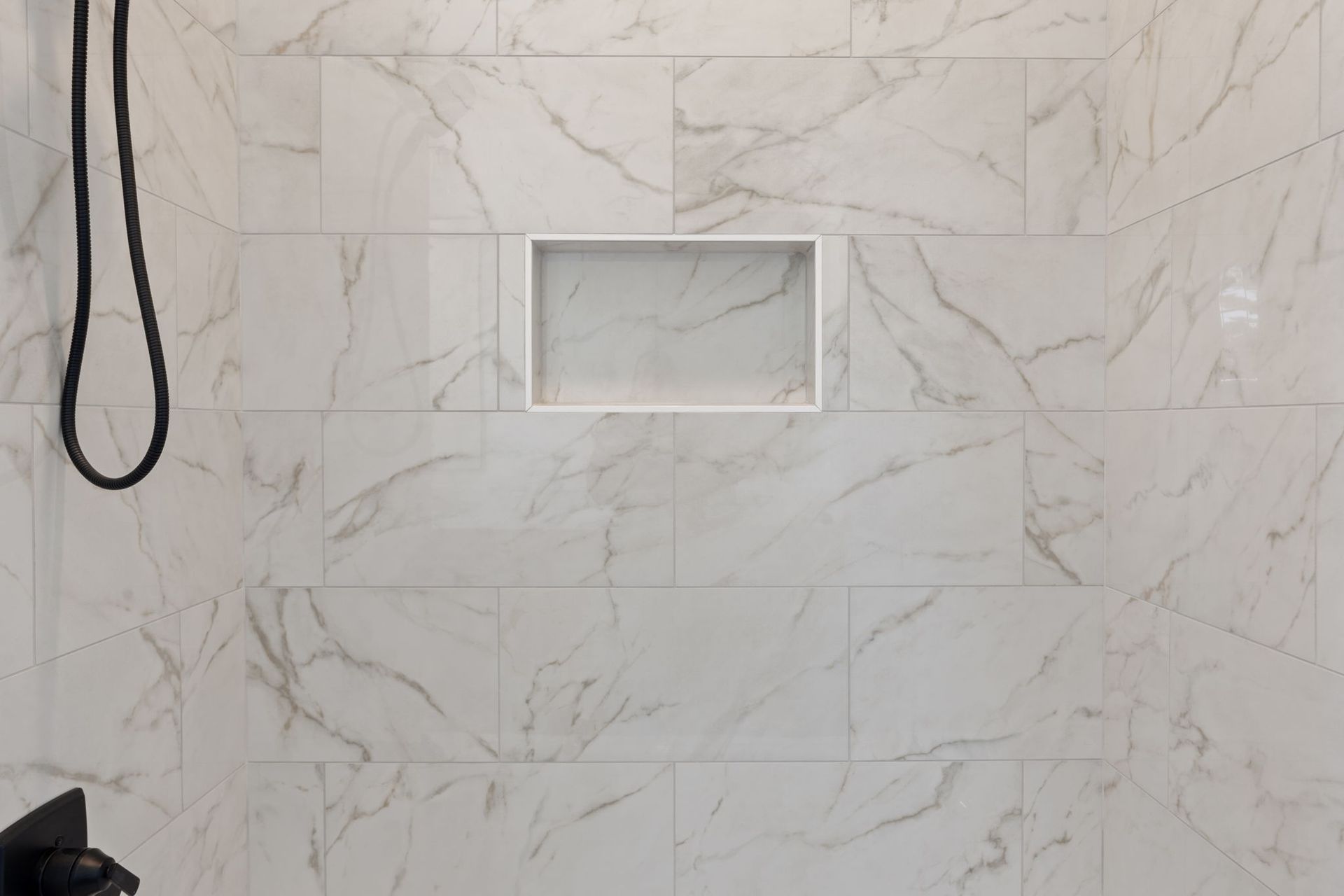 White marble-tiled shower wall with a recessed niche and hanging black shower hose on the left