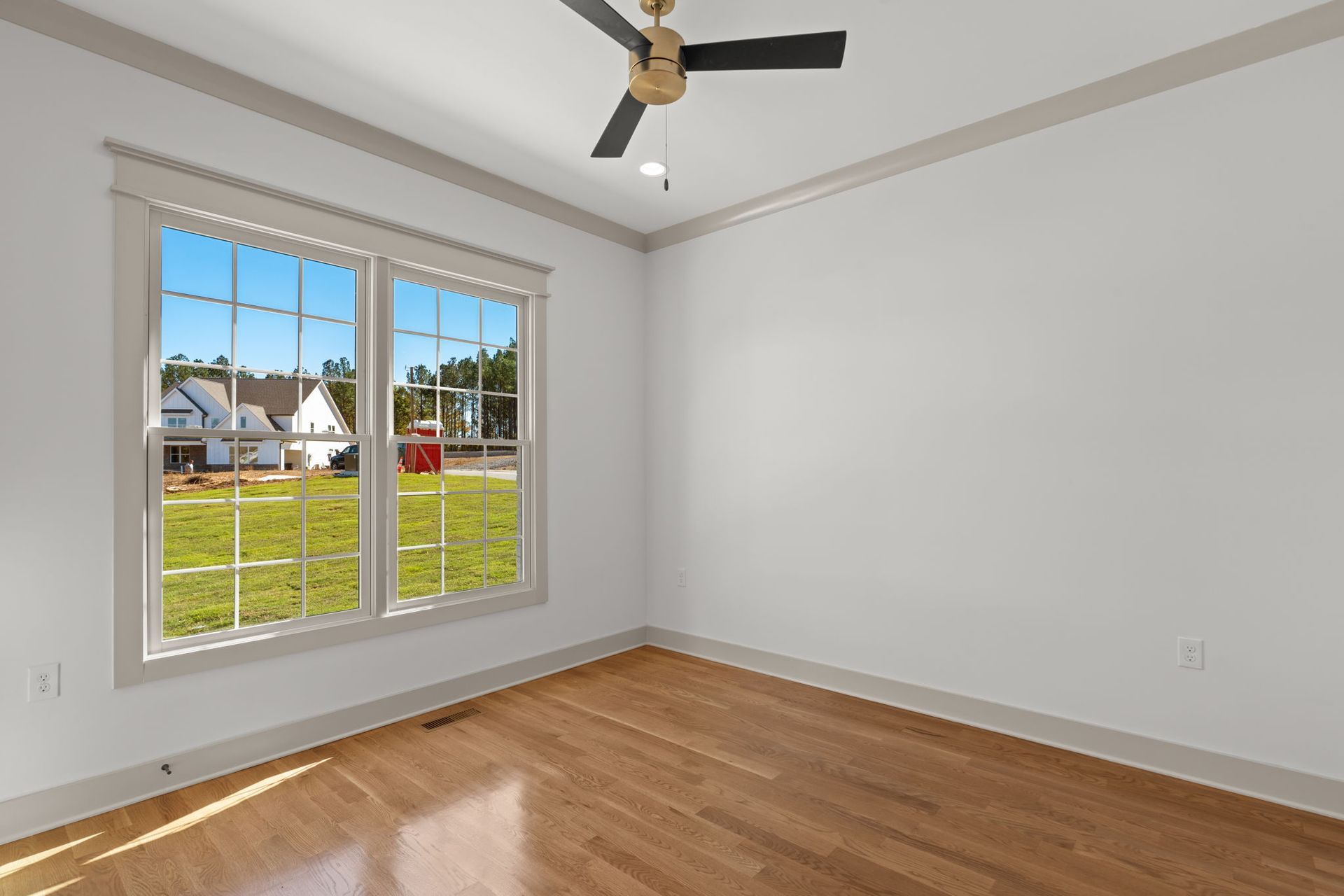 Empty room with hardwood floor, white walls, a ceiling fan, and a large window overlooking a yard.