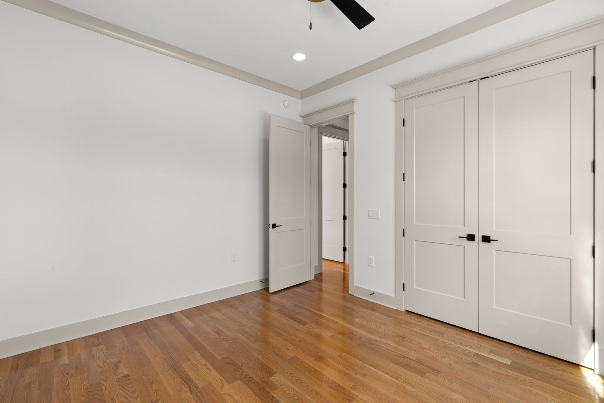 Empty white bedroom with hardwood floors, ceiling fan, open door, and double closet doors