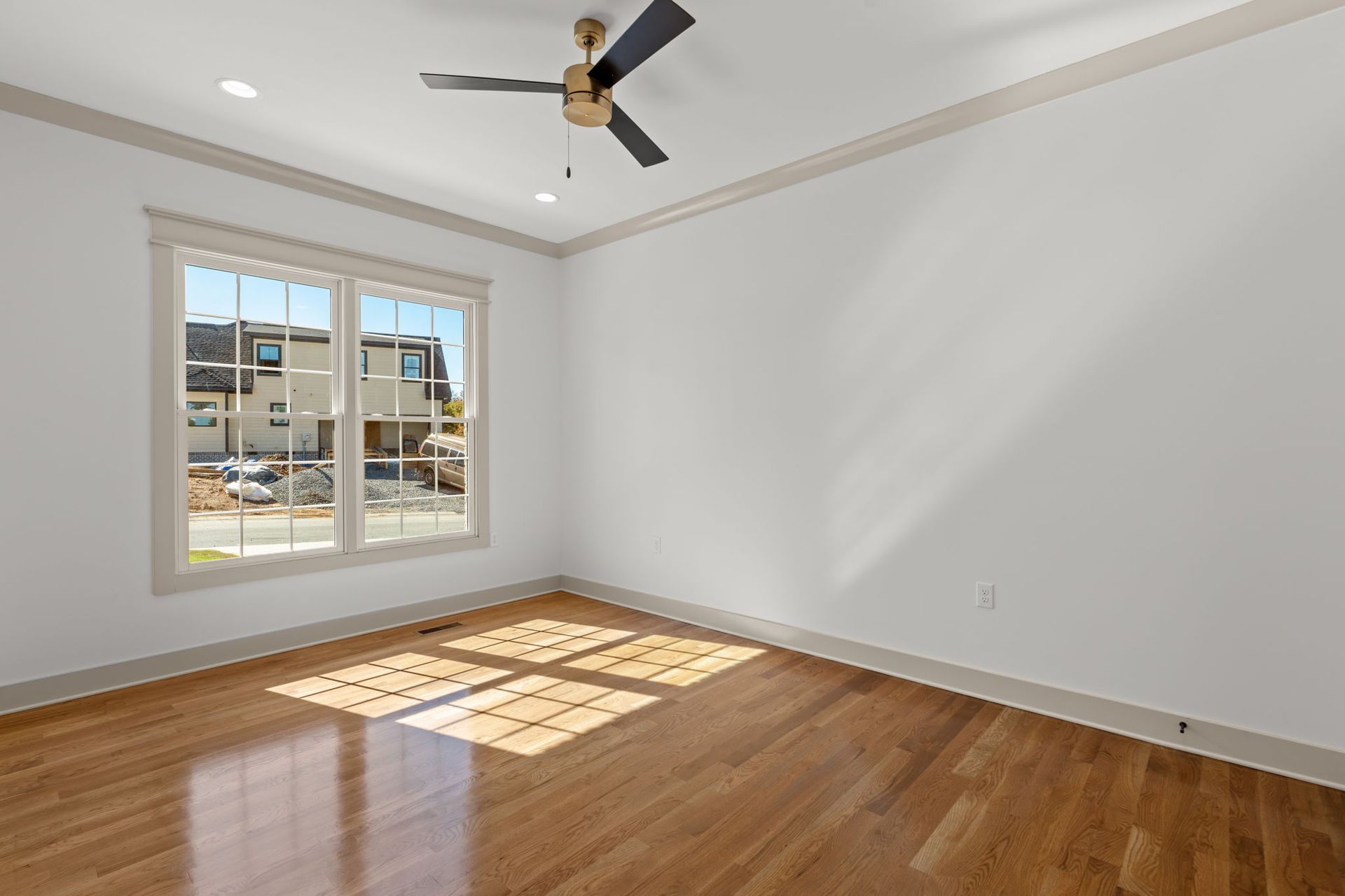 Empty sunlit room with hardwood floors, gray walls, ceiling fan, and a window casting light on the floor