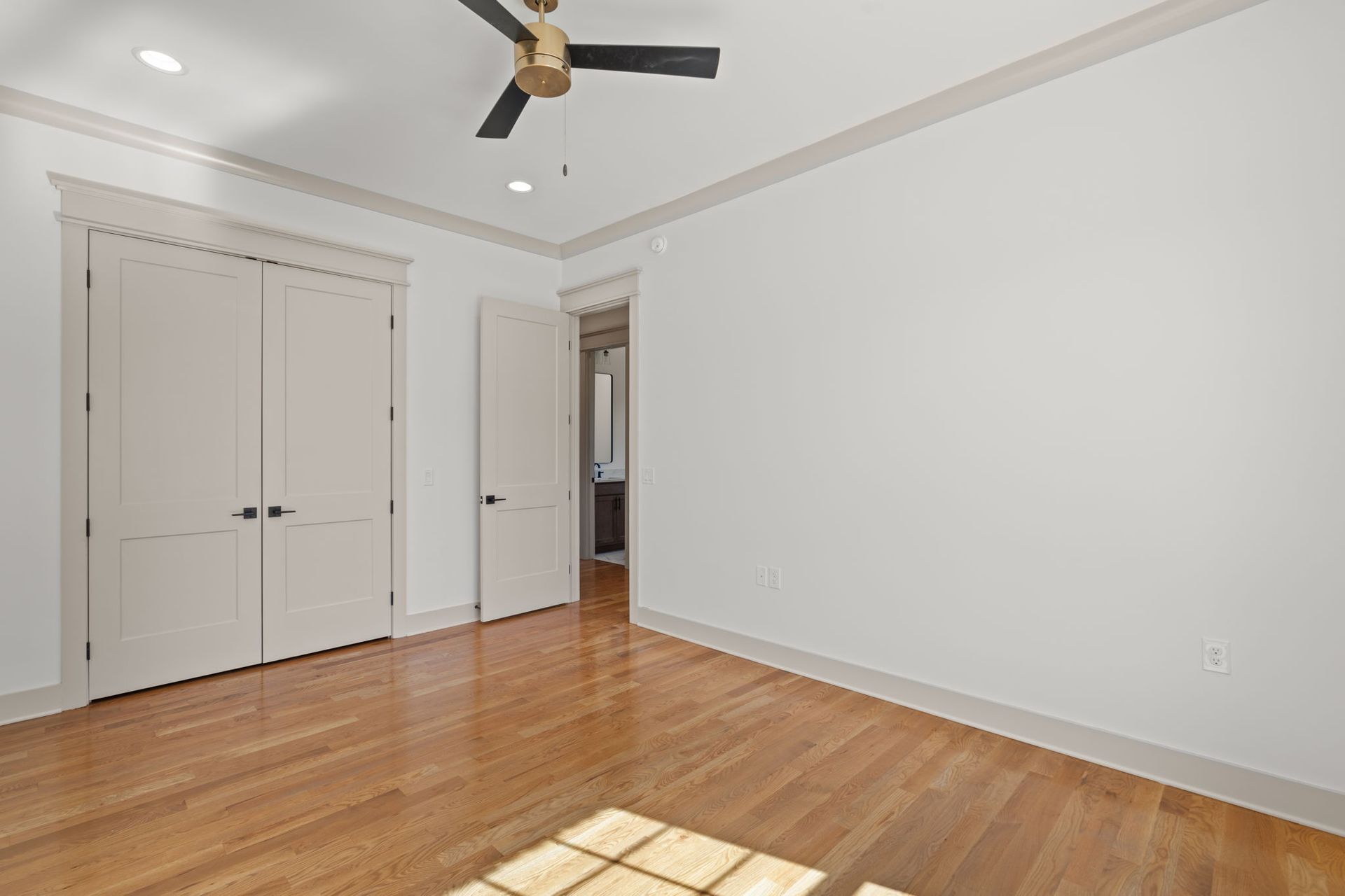 Empty white bedroom with hardwood floors, double closet doors, ceiling fan, and sunlight on the floor