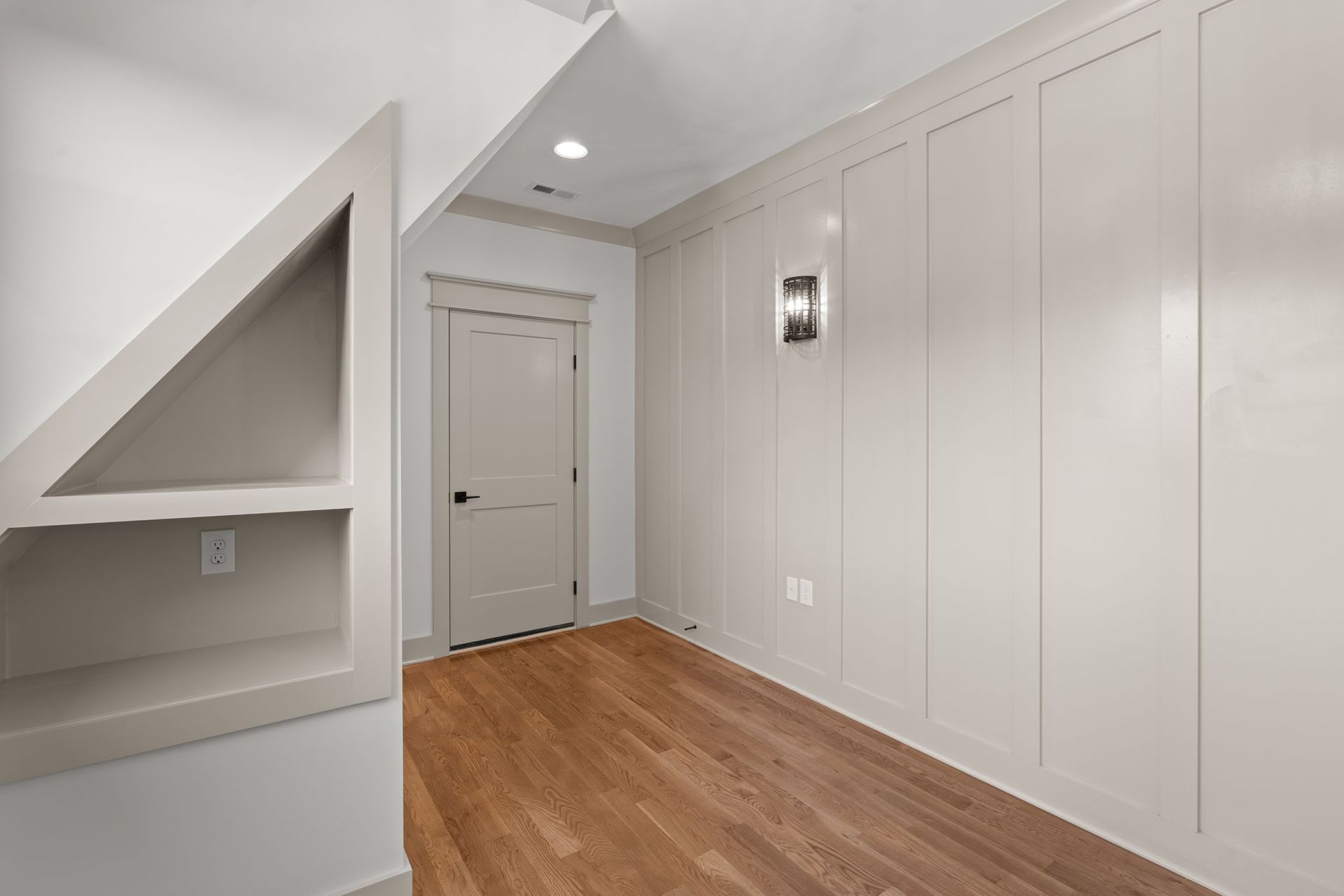 Bright hallway with white paneled walls, wood floor, and a gray door under a sloped ceiling.