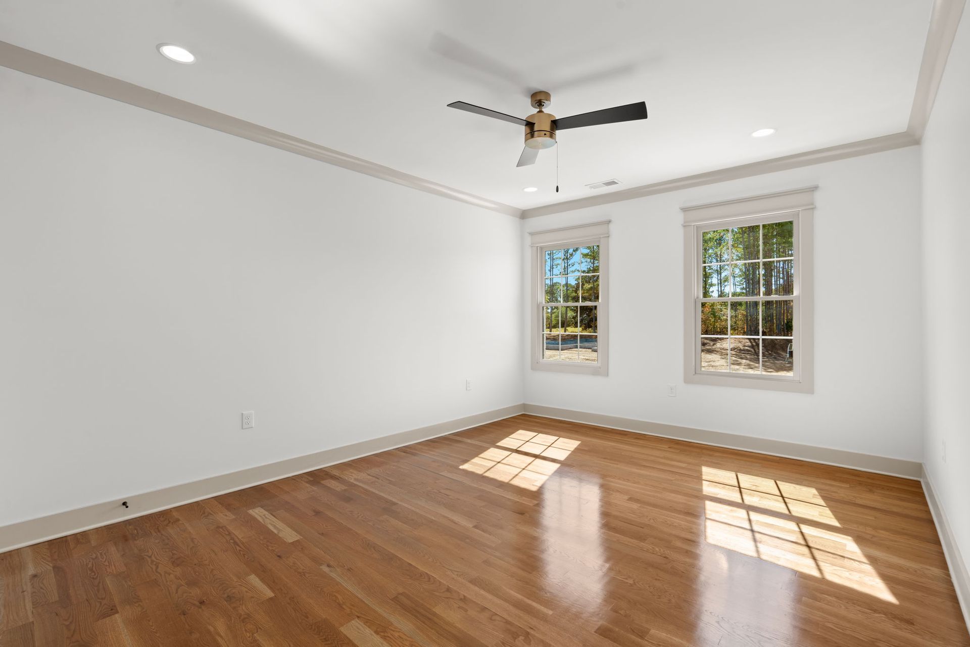 Empty white bedroom with hardwood floors, two windows, and a ceiling fan.