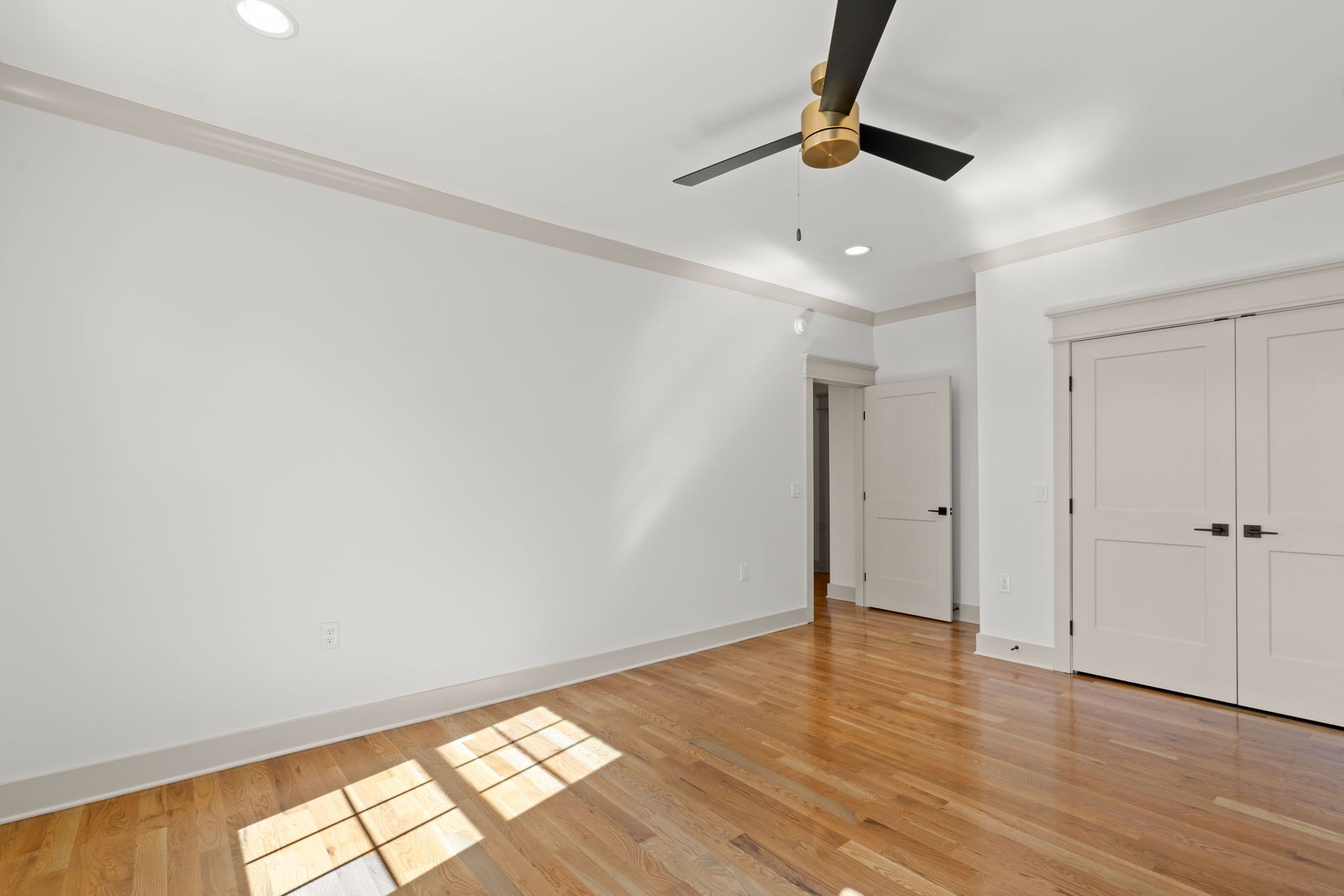 Empty white bedroom with hardwood floors, ceiling fan, and double closet doors with sunlight on the floor