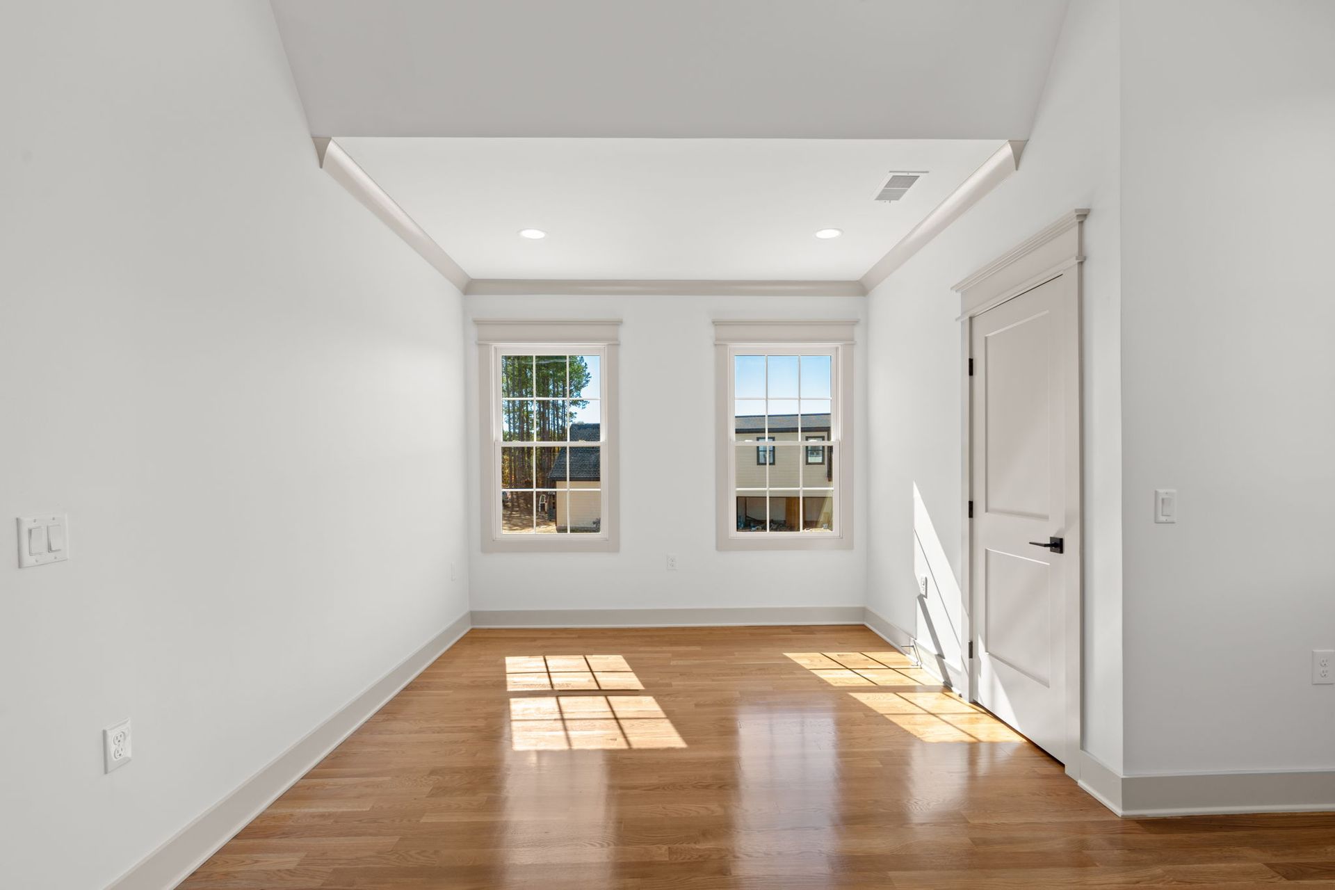 Bright empty room with white walls, wood floor, two windows, and a white door.
