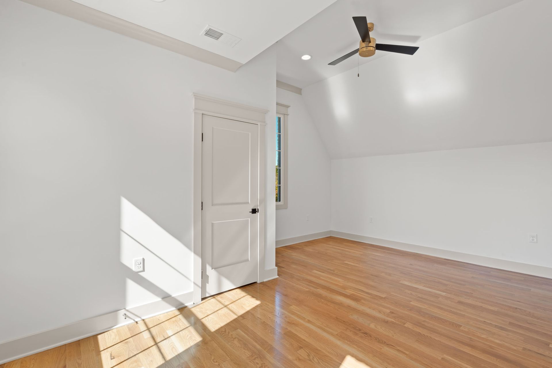 Empty white room with wood floor, slanted ceiling, door, and ceiling fan in bright sunlight