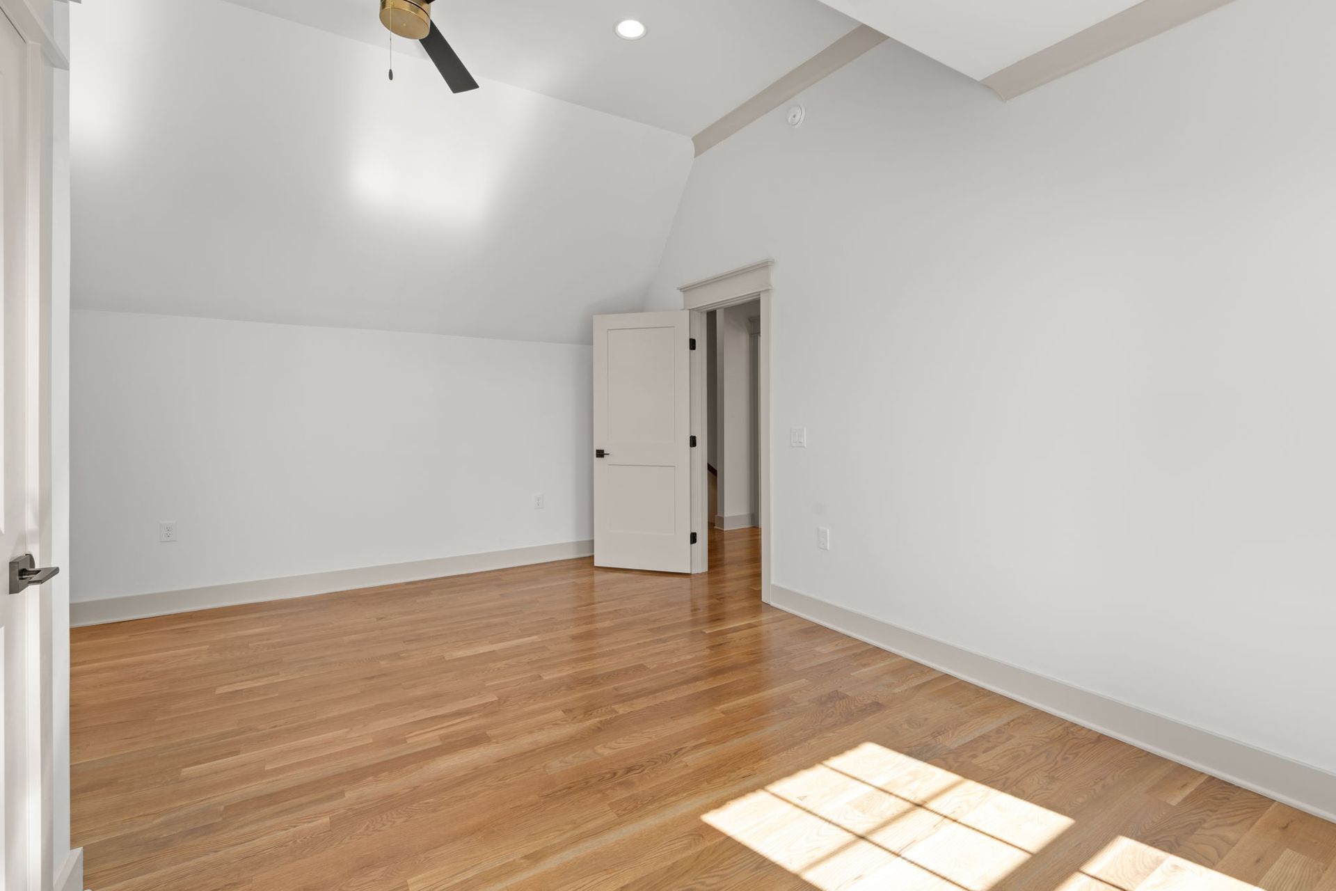Empty white bedroom with hardwood floors and a sloped ceiling, sunlight on the floor.
