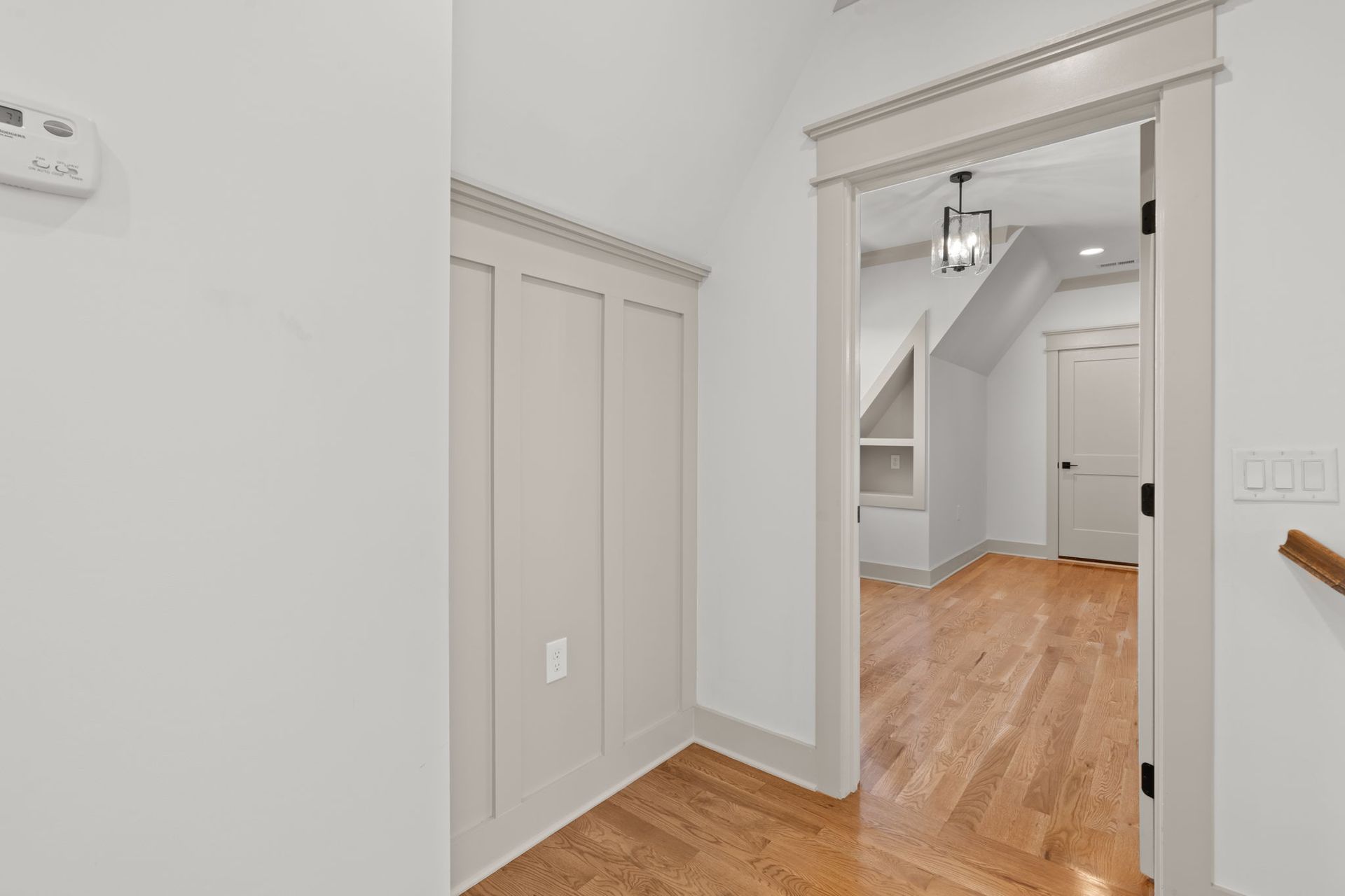 Empty white hallway with hardwood floors, closet doors, and an open doorway to a room with stairs and a light fixture