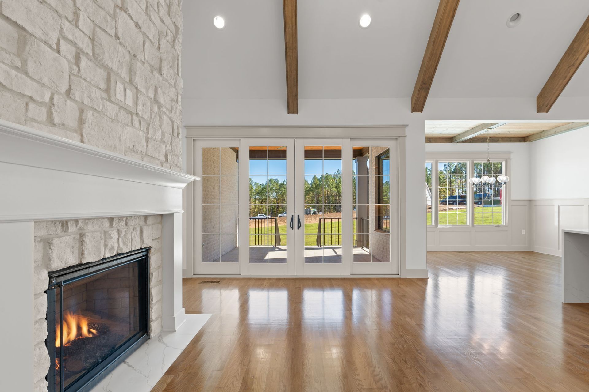 Bright living room with stone fireplace, hardwood floors, exposed beams, and large windows overlooking a yard
