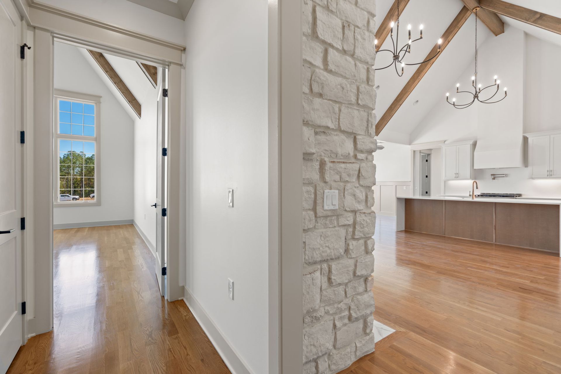 Bright hallway with wood floors, stone column, and view into a vaulted kitchen with white walls and island.