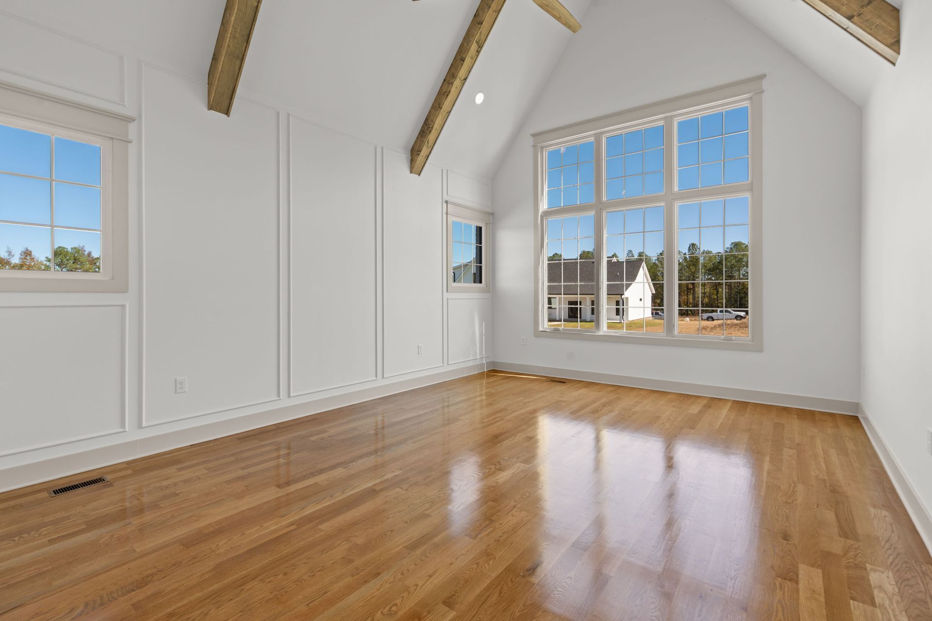 Bright empty room with hardwood floors, white walls, exposed beams, and large windows.