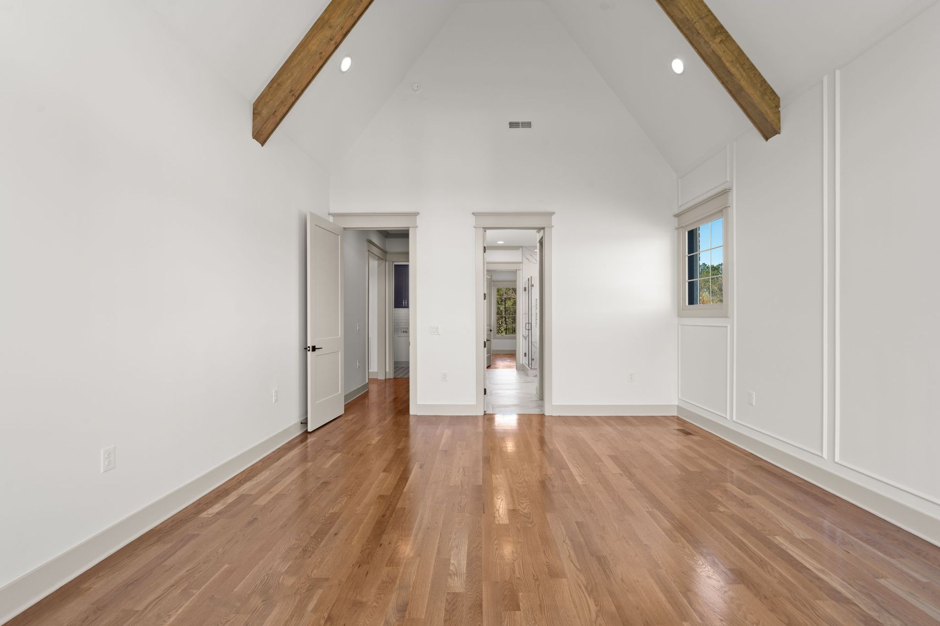 Empty white room with vaulted wood-beam ceiling and polished hardwood floors, seen through an open doorway.