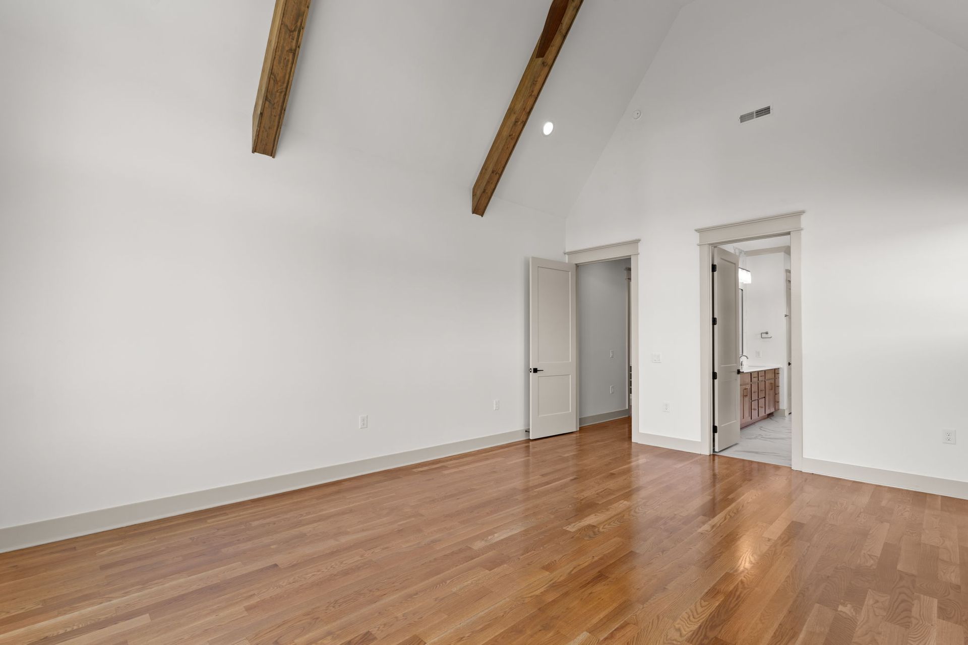 Empty white room with hardwood floors, exposed beams, and two open doors.