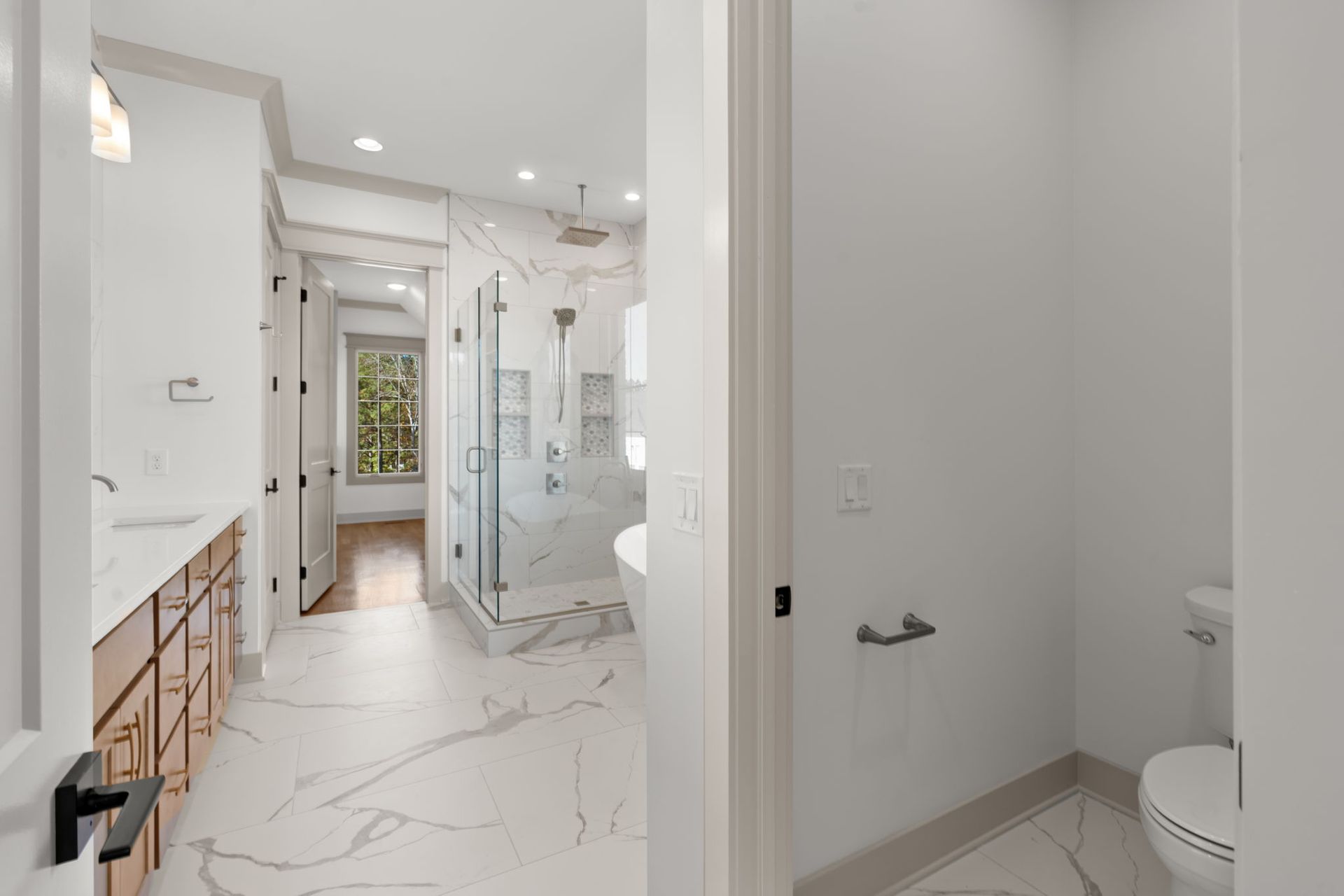 Bright modern bathroom with white marble tile, glass shower, and a window at the far end.