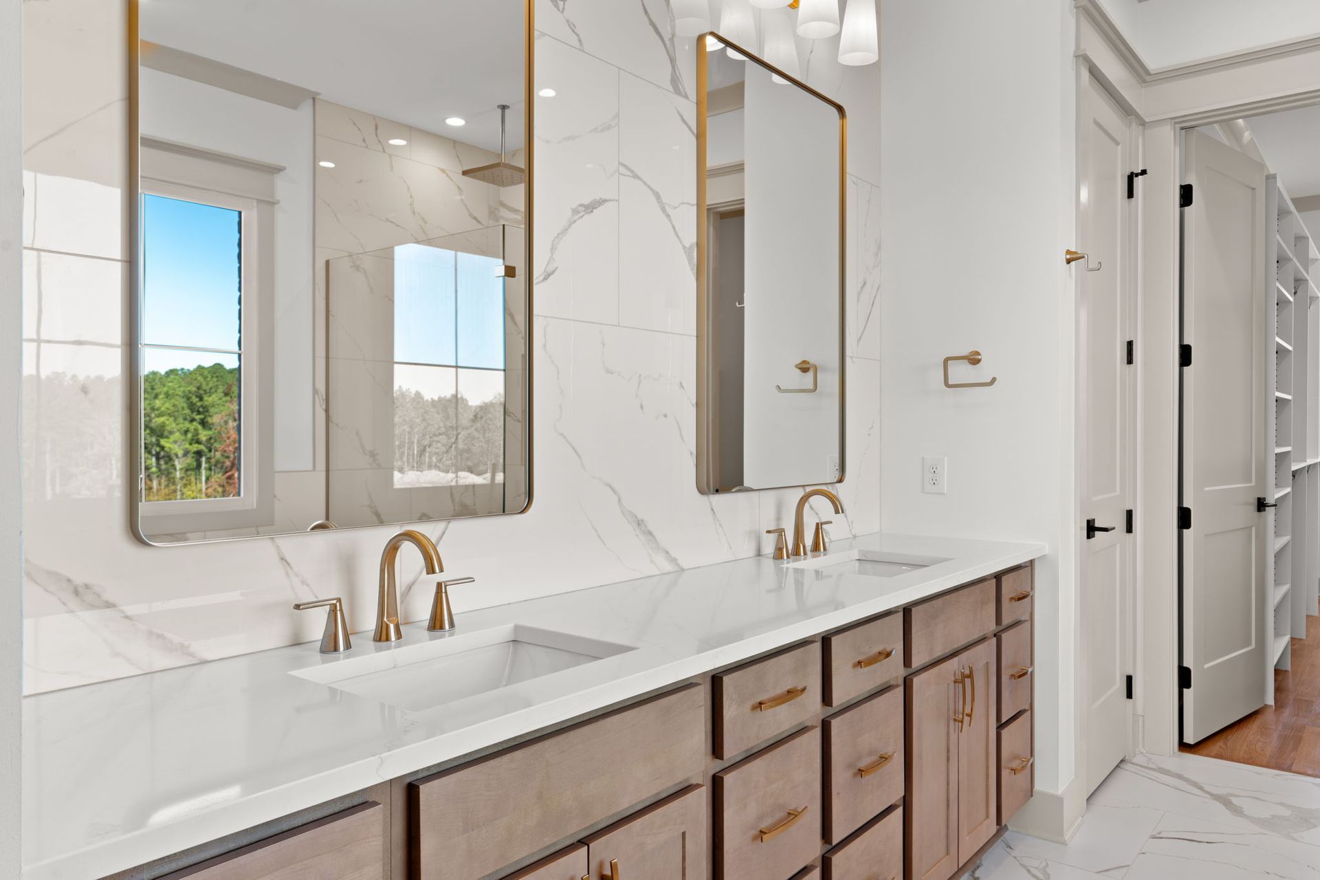 Modern white bathroom with double vanity, gold fixtures, marble walls, and large mirrors.