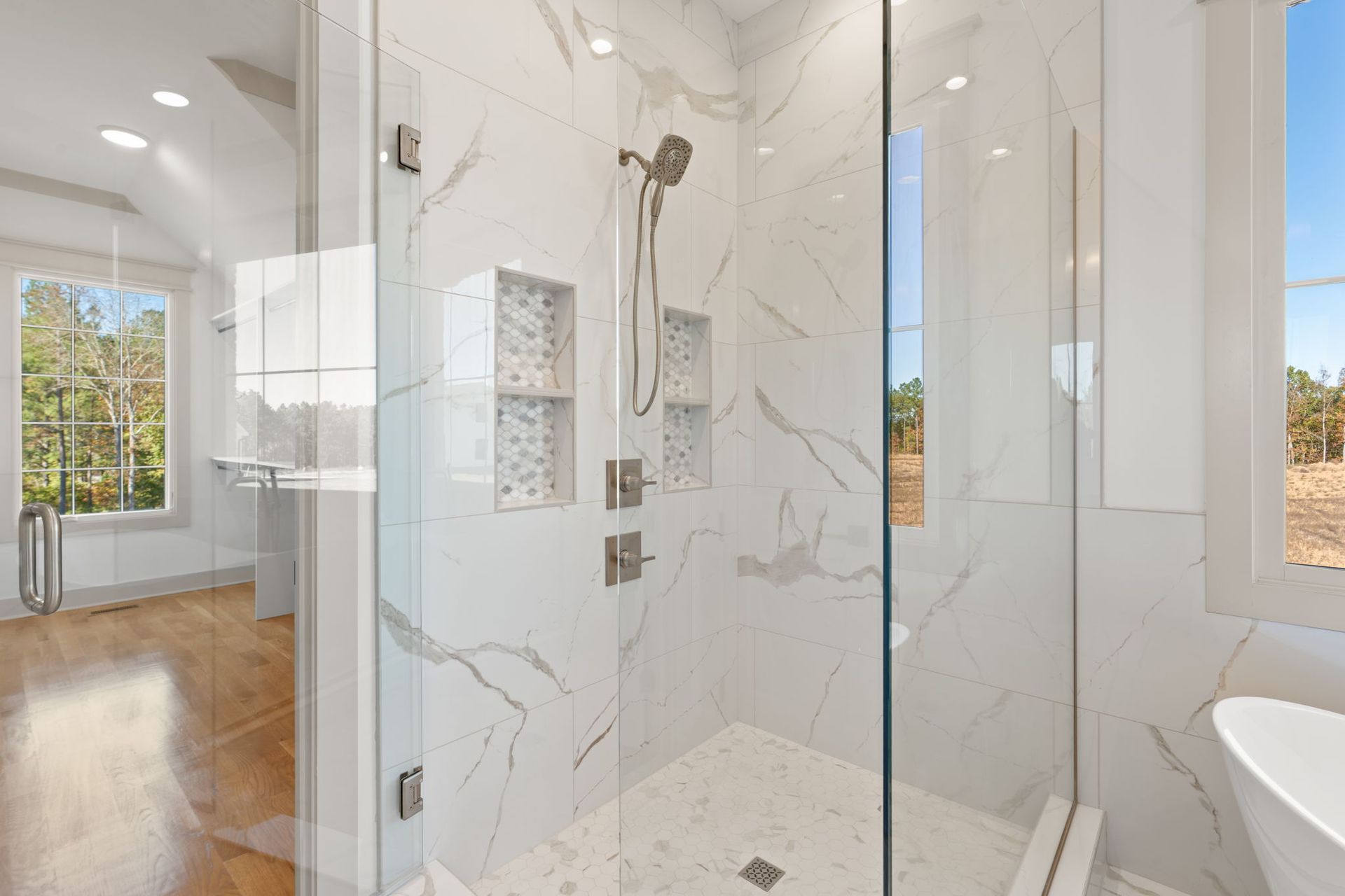 Bright modern bathroom with glass shower, white marble tile, and large windows overlooking trees