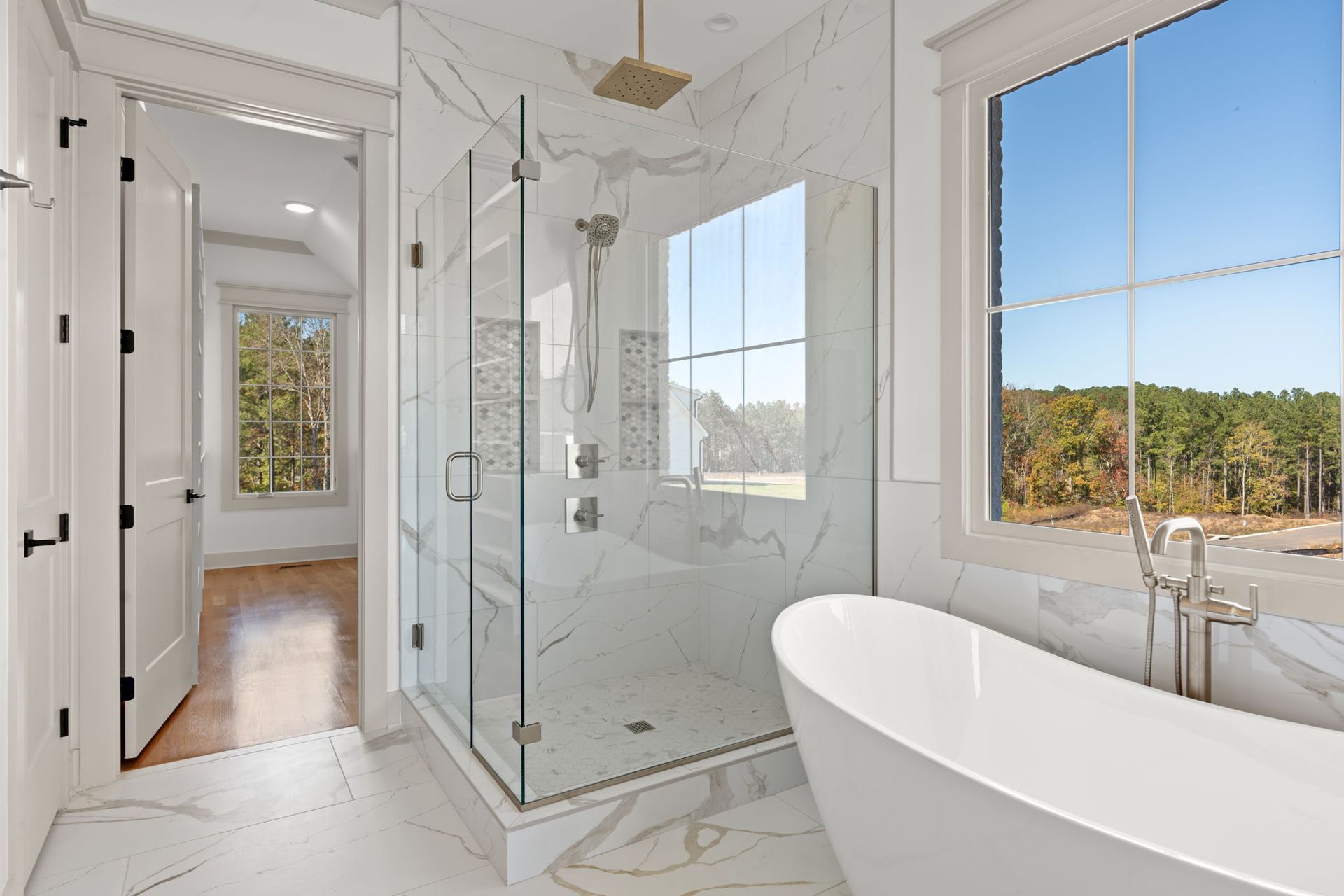 Bright white bathroom with glass shower, freestanding tub, and large window overlooking trees
