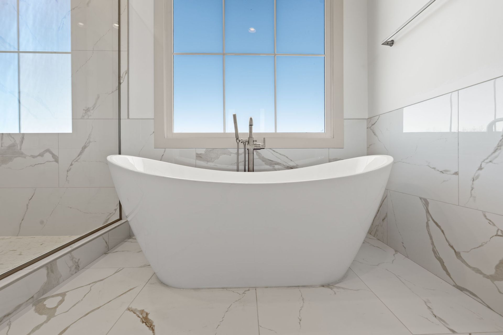 Modern white bathroom with freestanding tub, marble walls, and a large window.