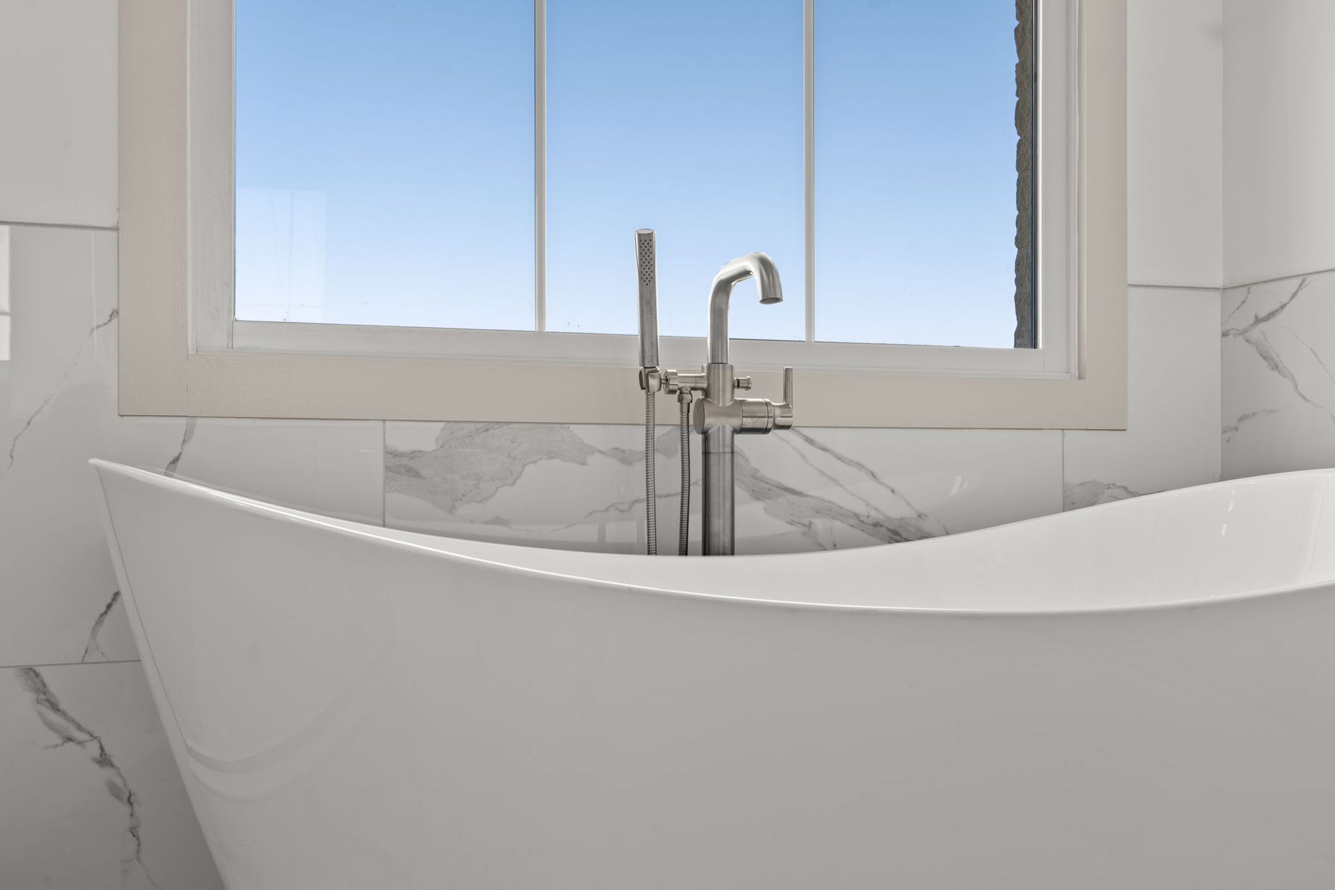Modern white bathroom with a freestanding tub under a window and chrome faucet