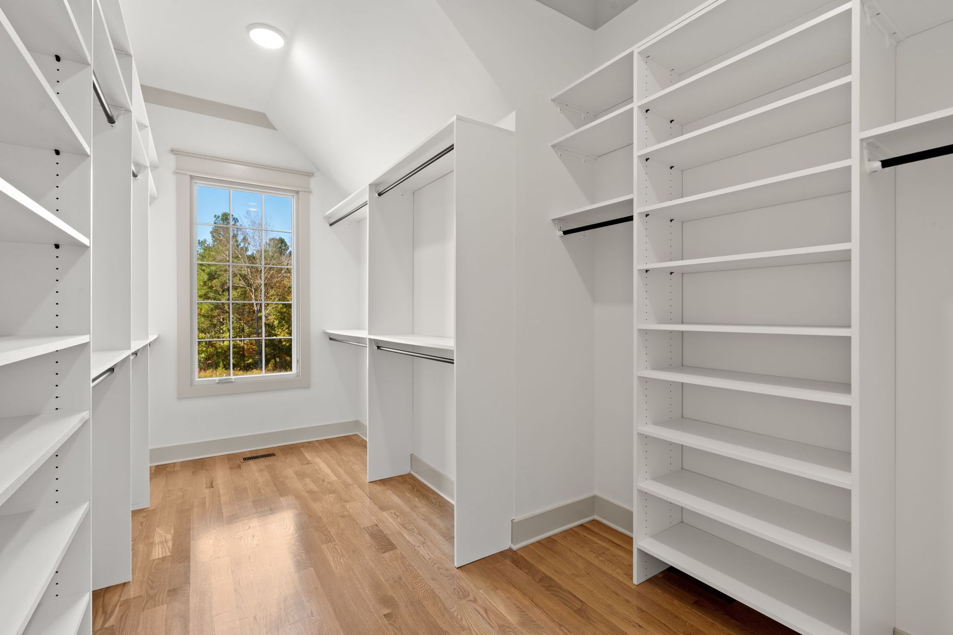 Bright white walk-in closet with built-in shelves, hanging rods, and wood floors by a window