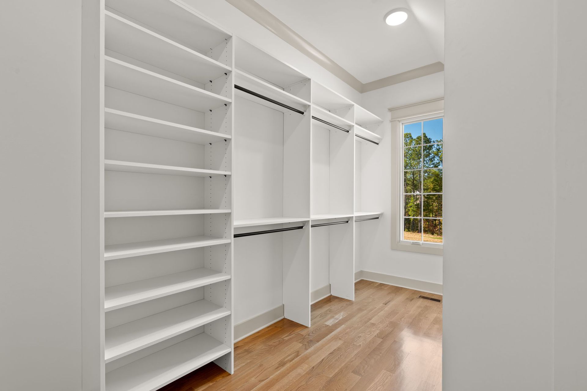Bright white walk-in closet with built-in shelves, hanging rods, and a window over wood floors