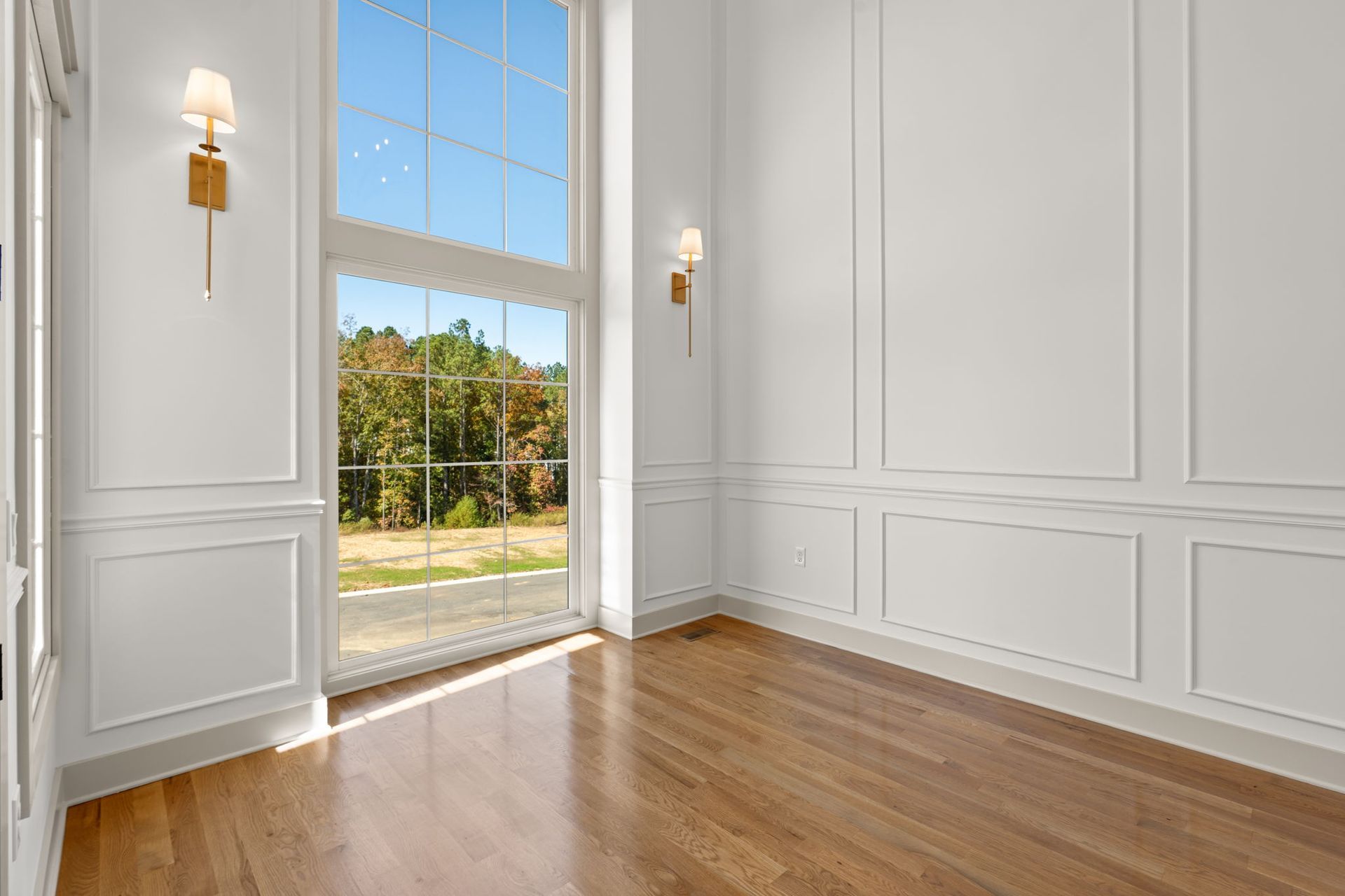 Bright empty room with white paneled walls, hardwood floor, wall sconces, and a tall glass door to a sunny view