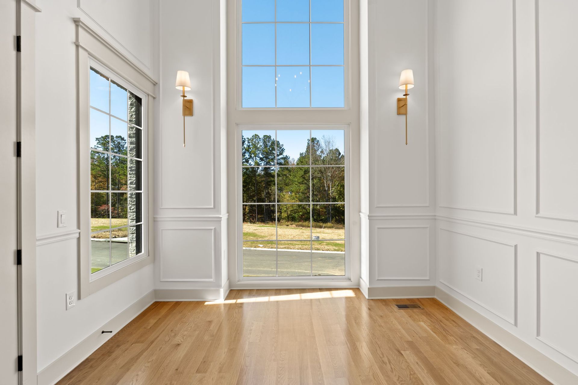 Bright empty room with hardwood floors, white walls, large windows, and glass door overlooking trees and grass