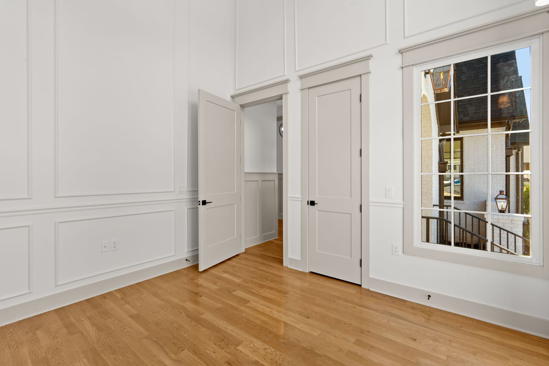 Bright empty room with white paneled walls, open door, hardwood floor, and tall window.