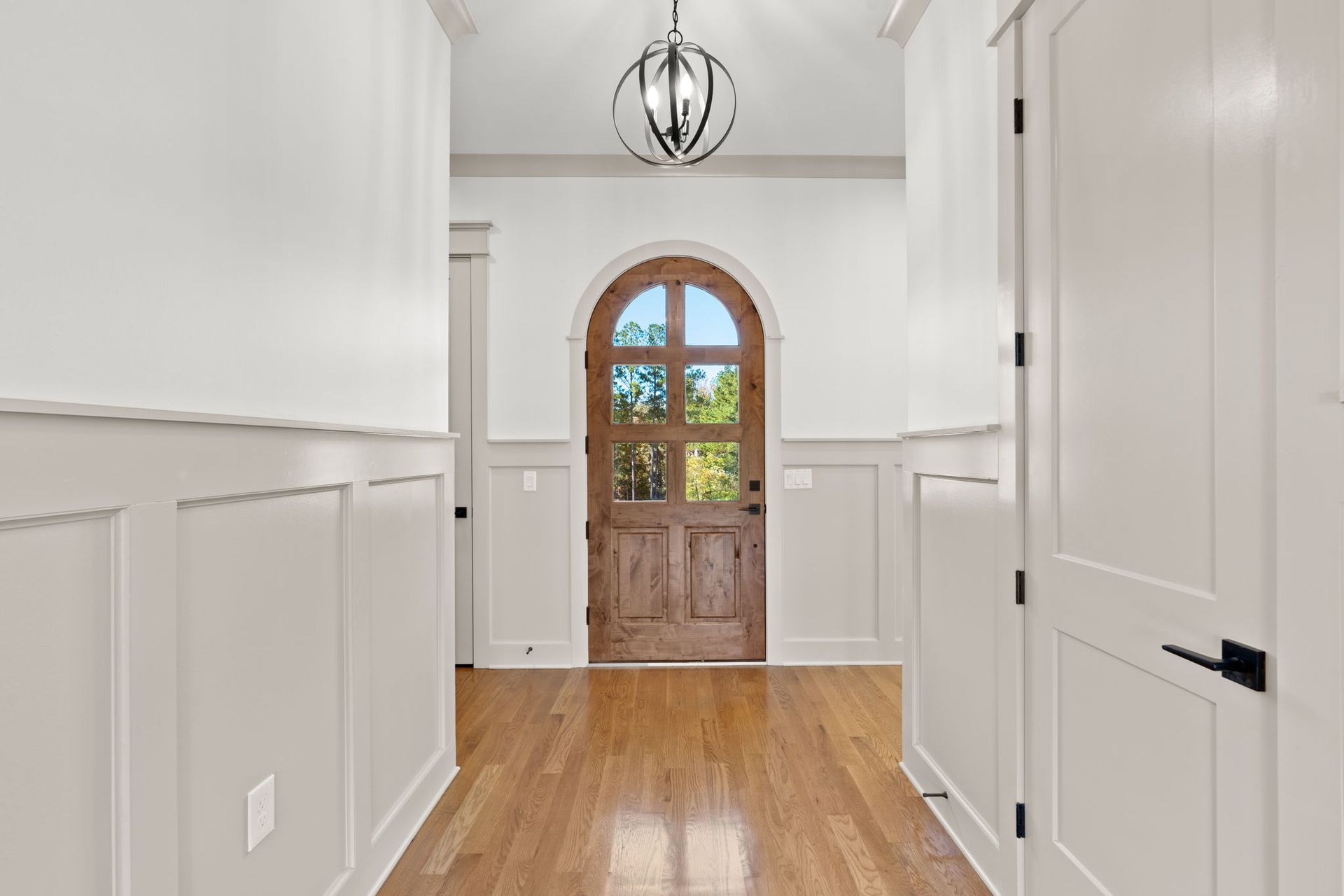 Bright white entry hallway with wood floor, wainscoting, pendant light, and an arched wooden front door.