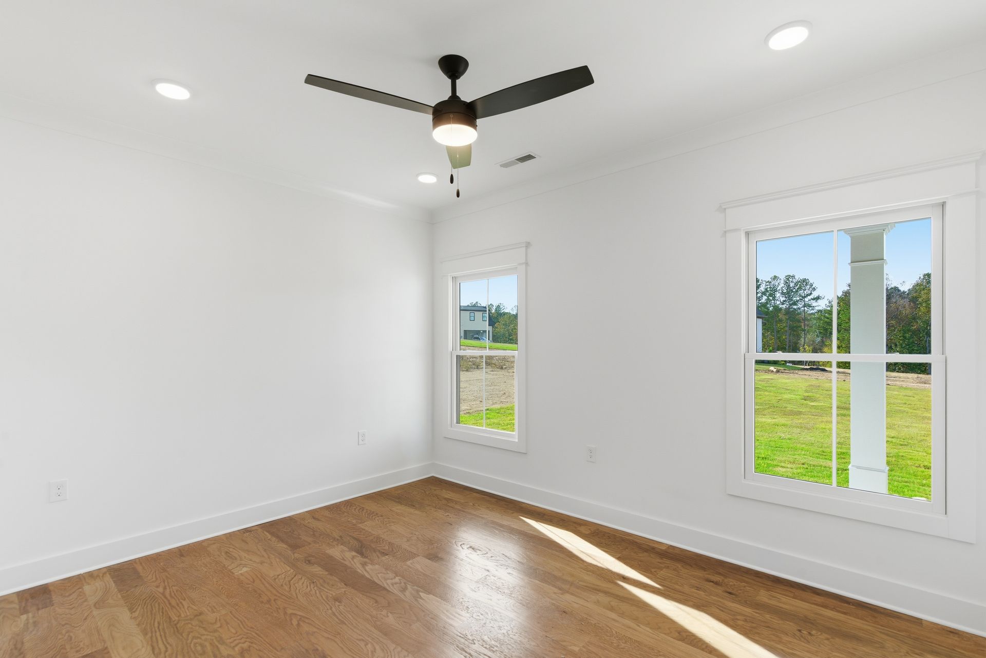 Empty white room with hardwood floors, ceiling fan, and two windows overlooking a green yard