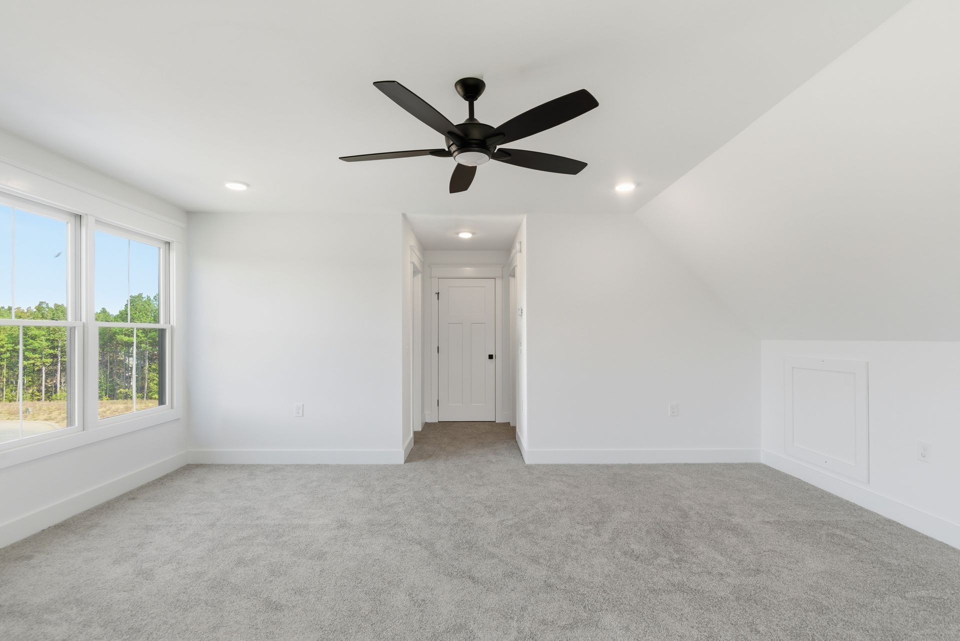Bright empty bedroom with gray carpet, white walls, ceiling fan, and a window view.