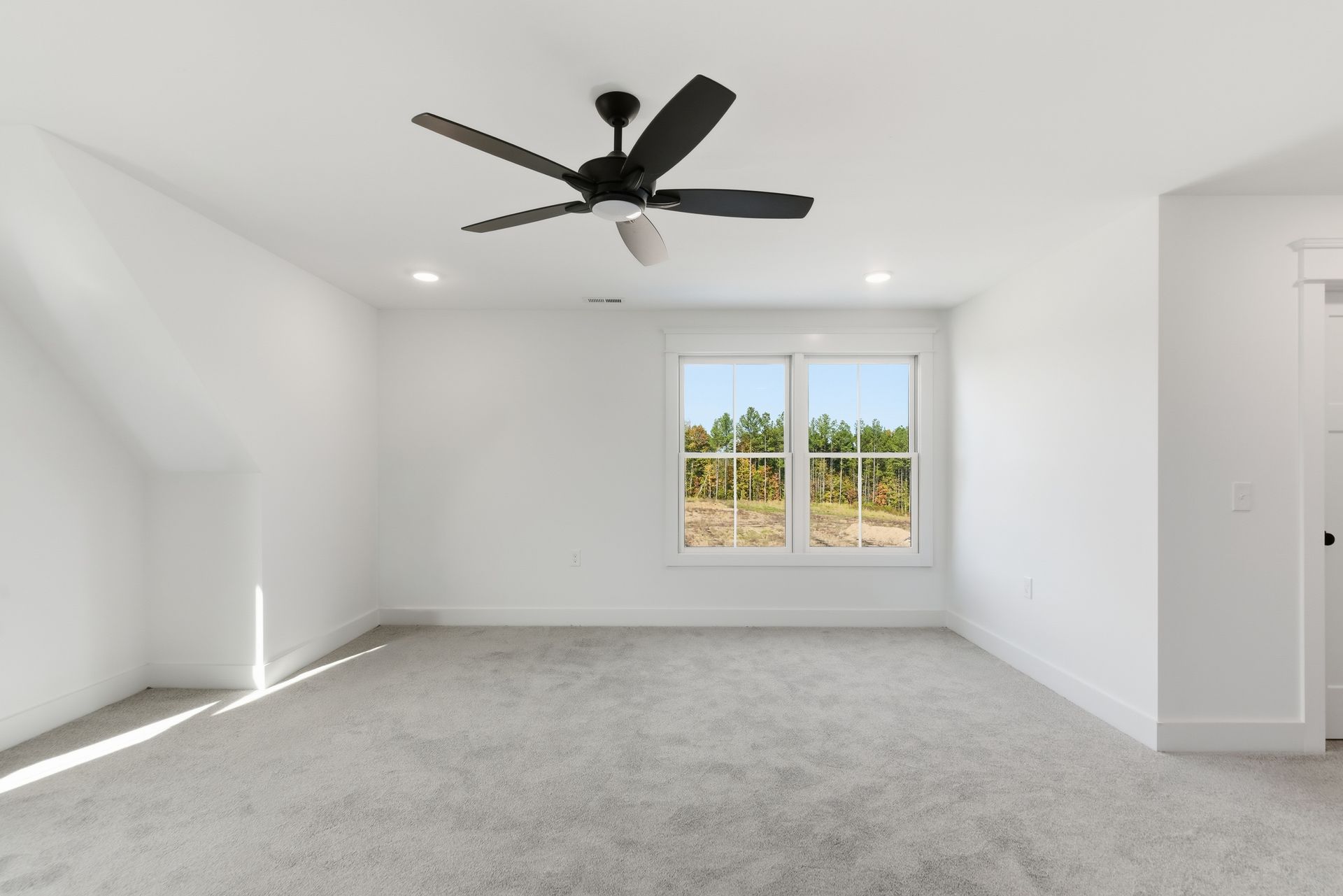Empty white bedroom with gray carpet, ceiling fan, and window view of trees and field