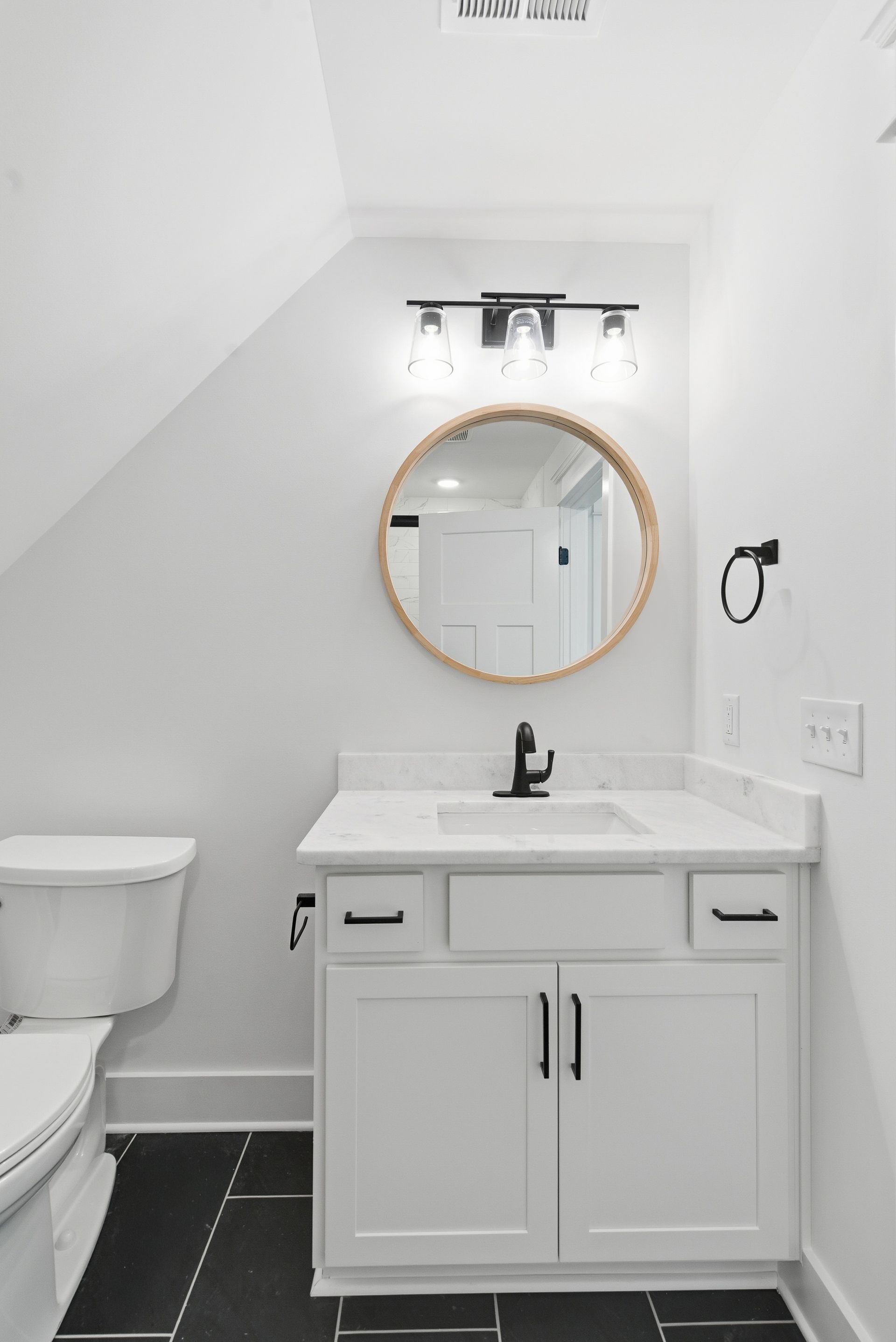 Small white bathroom with vanity, round mirror, toilet, and black tile floor.