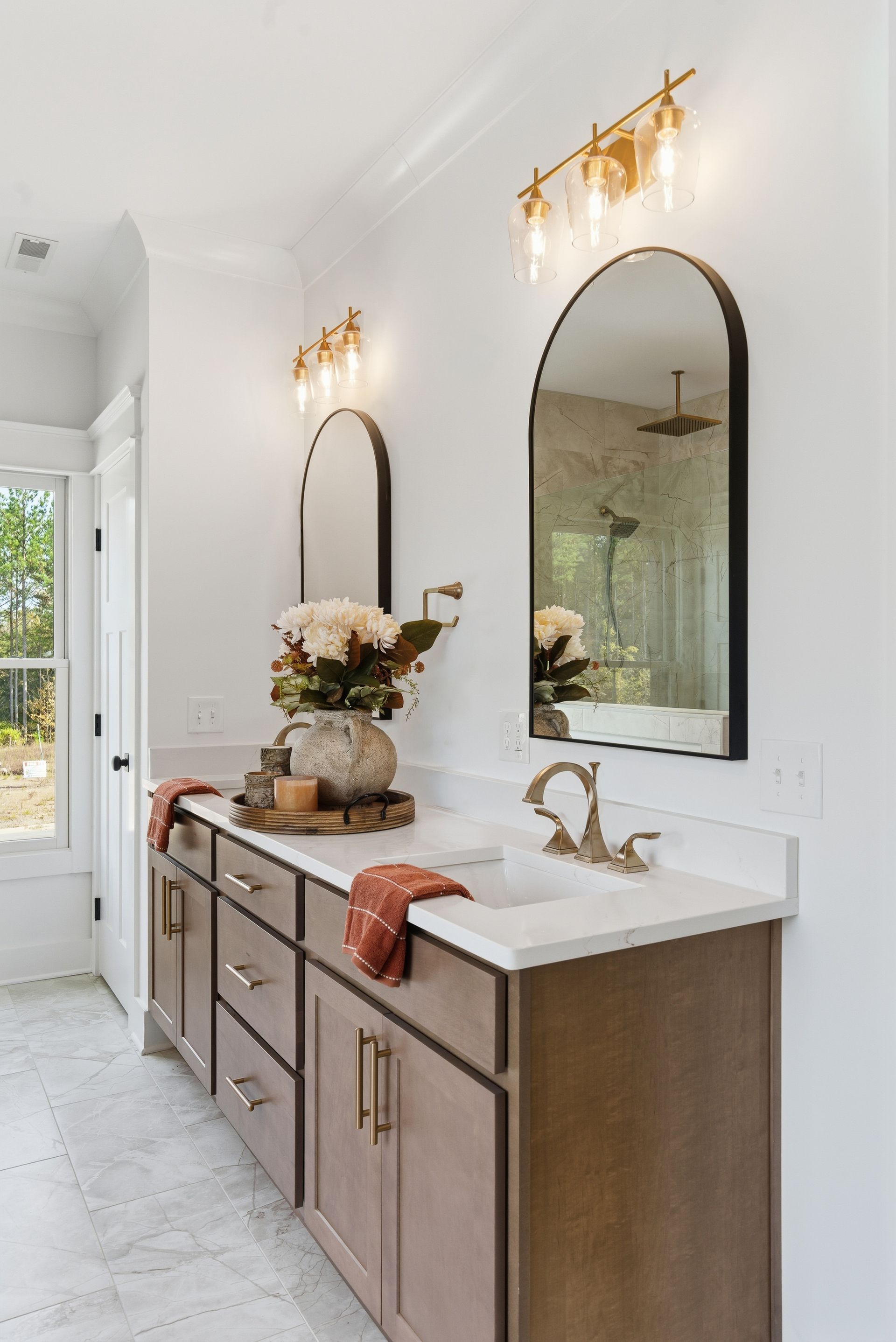 Elegant bathroom vanity with white countertop, wood cabinets, gold fixtures, and arched mirrors under wall sconces