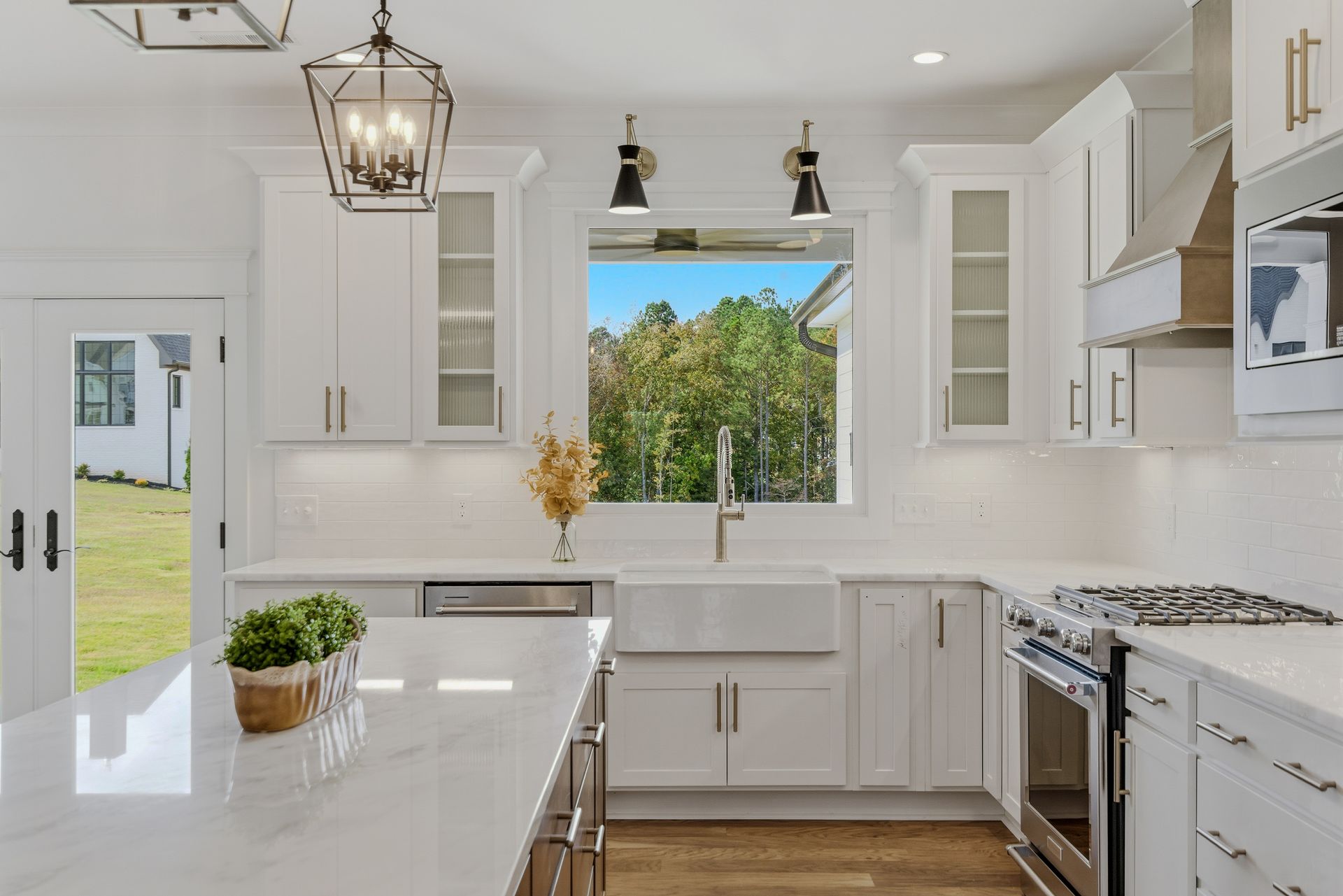 Bright white modern kitchen with island, stainless stove, and large window view of greenery.