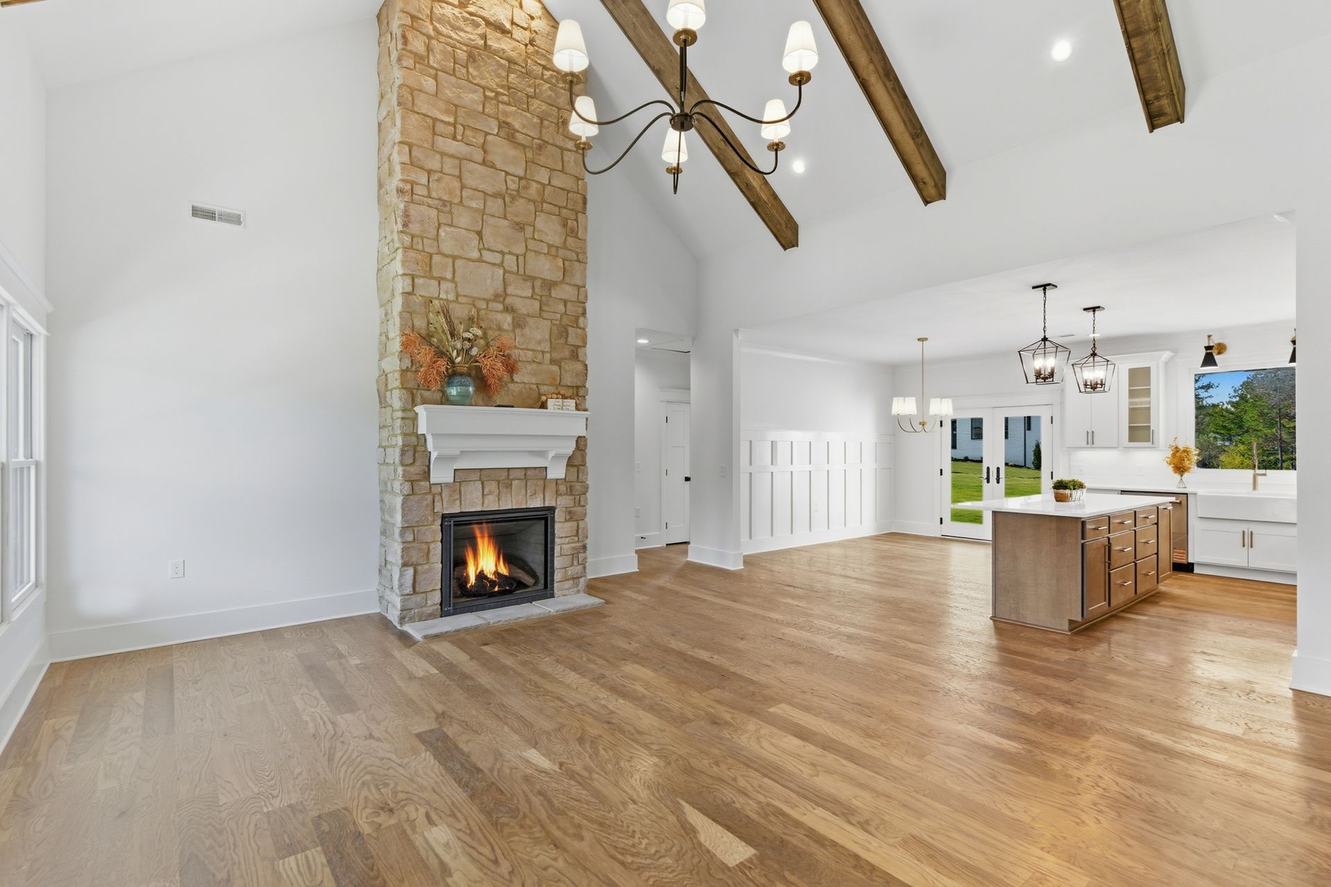 Bright open living room with stone fireplace, wood floors, exposed beams, and adjacent kitchen island