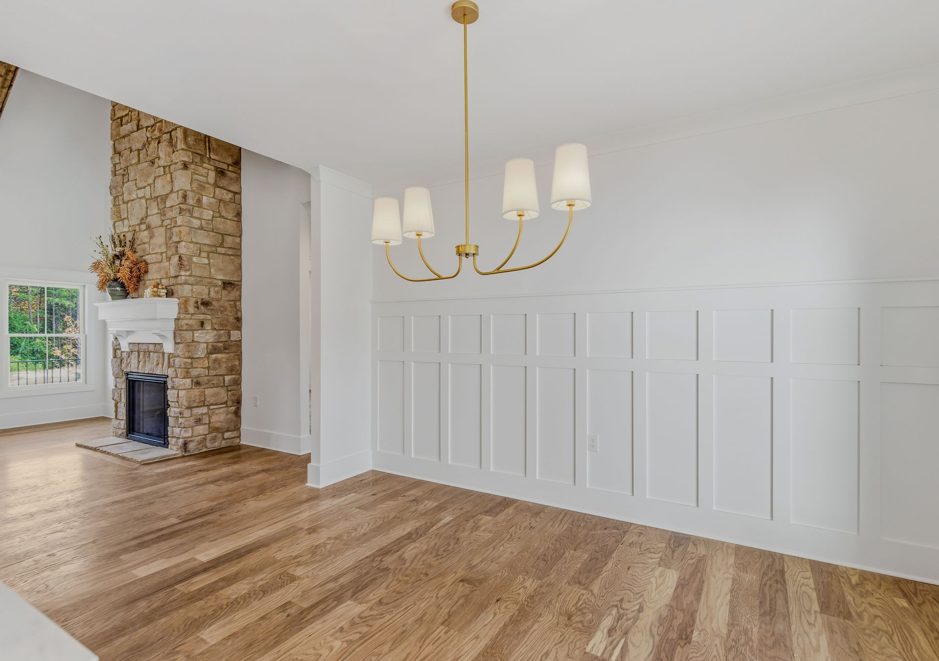 Bright empty dining room with white wainscoting, wood floors, and a stone fireplace.