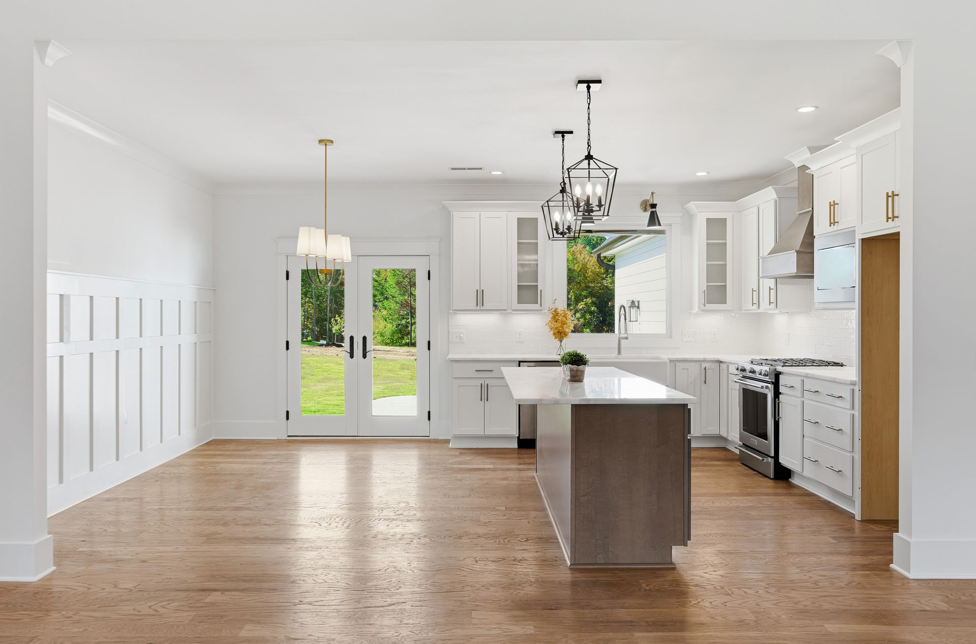 Bright, empty white kitchen with hardwood floors, island, white cabinets, and French doors to a green yard
