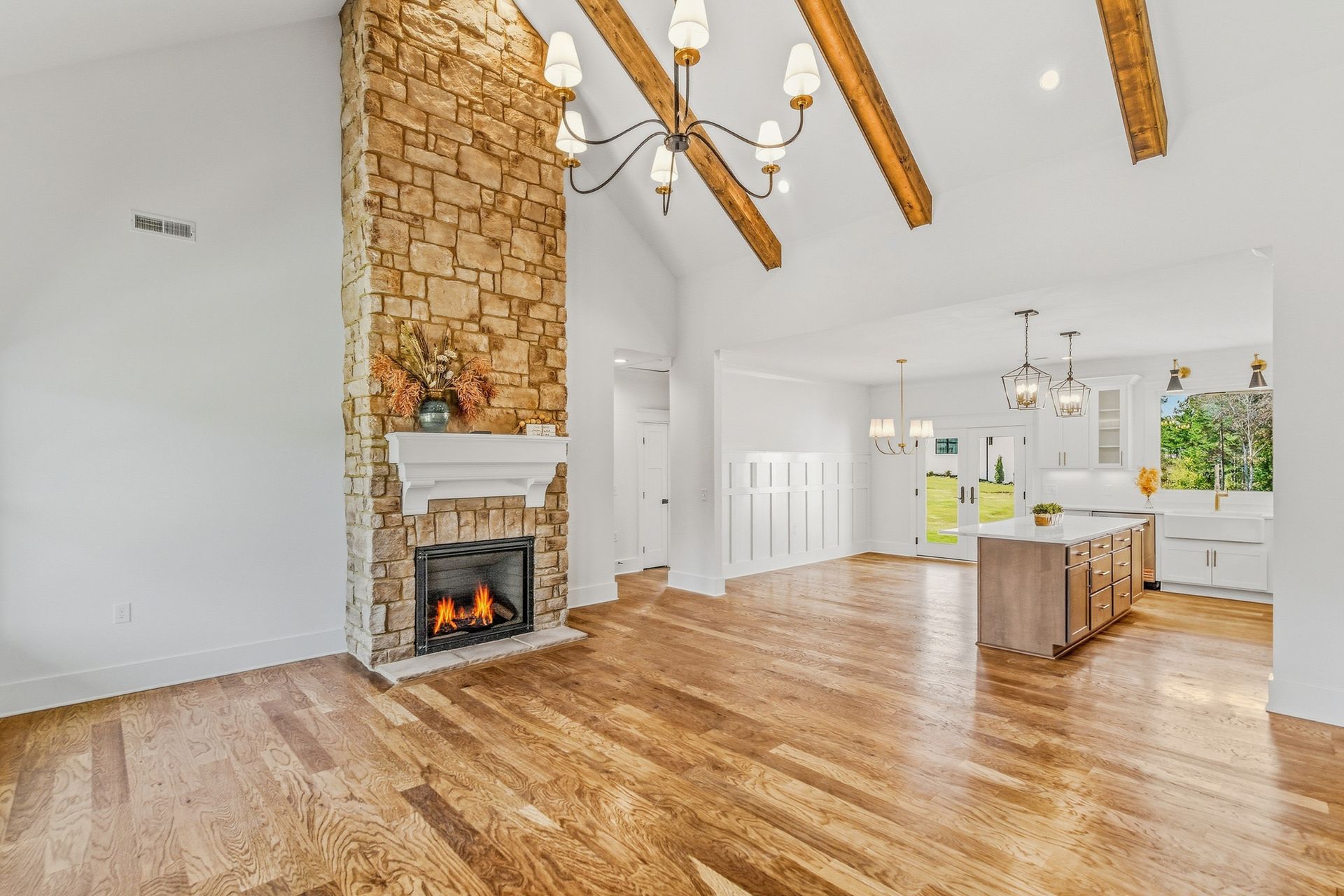Bright open living room with wood floors, stone fireplace, vaulted beams, and a white kitchen beyond.