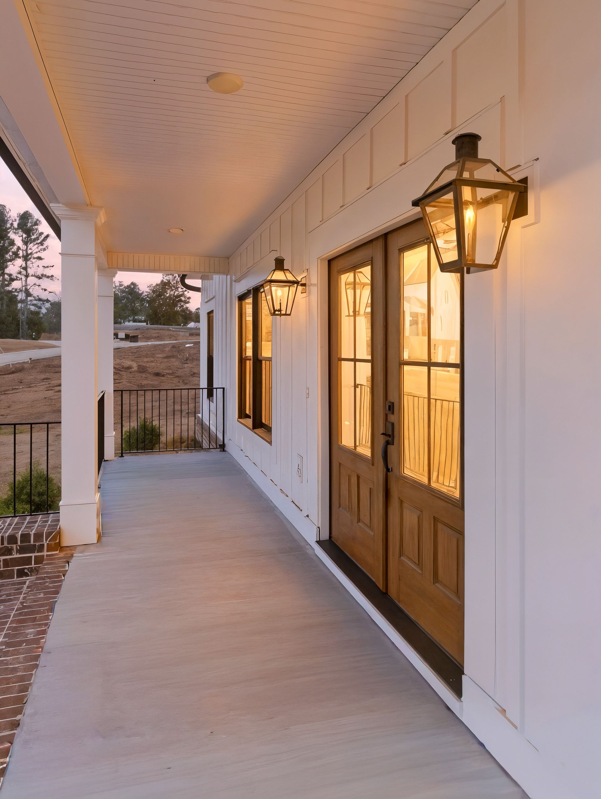 Covered front porch with white siding, hanging lanterns, and glass-paneled double doors at sunset